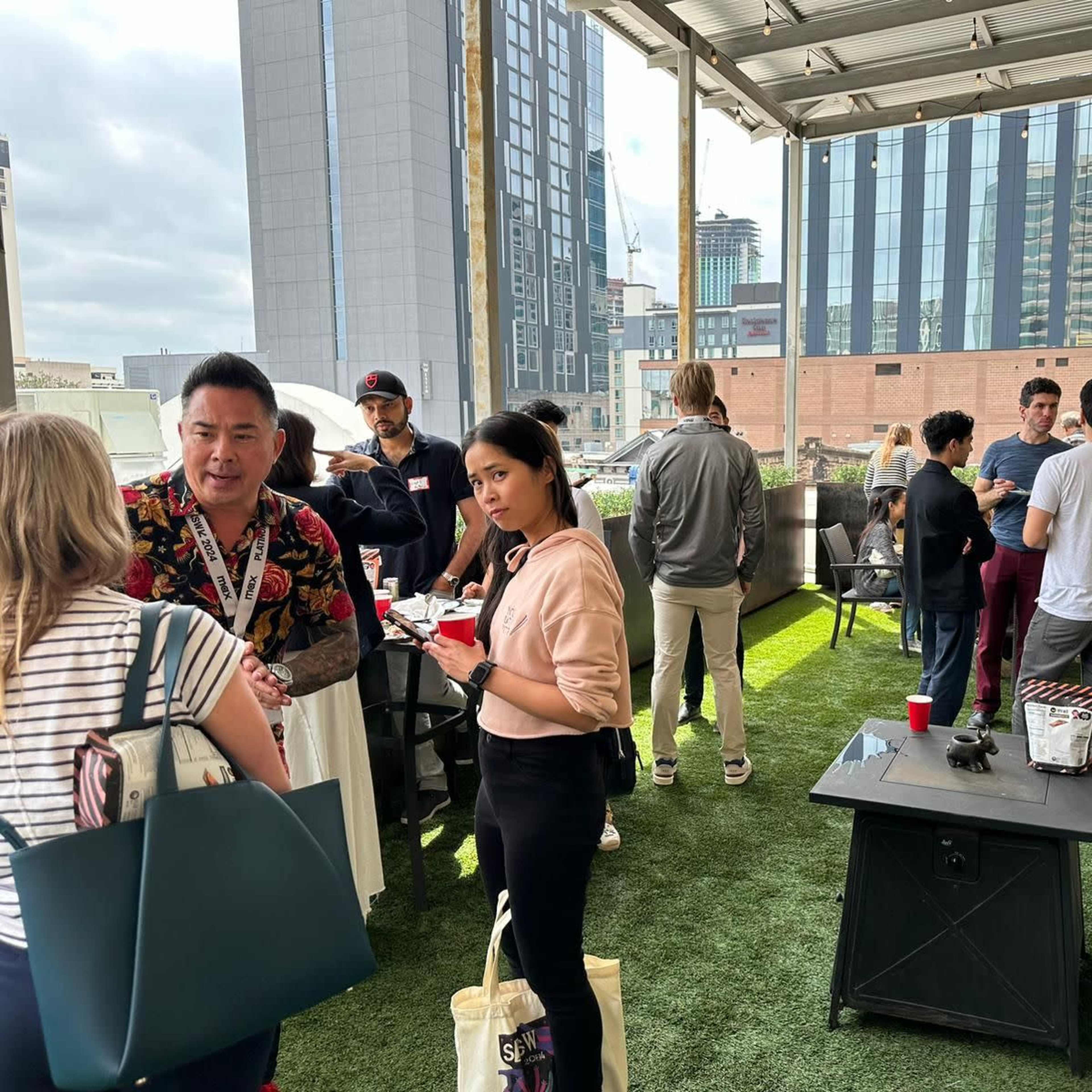 A group of people gathers on a rooftop patio, engaging in conversation and enjoying food and drinks against a backdrop of city buildings.