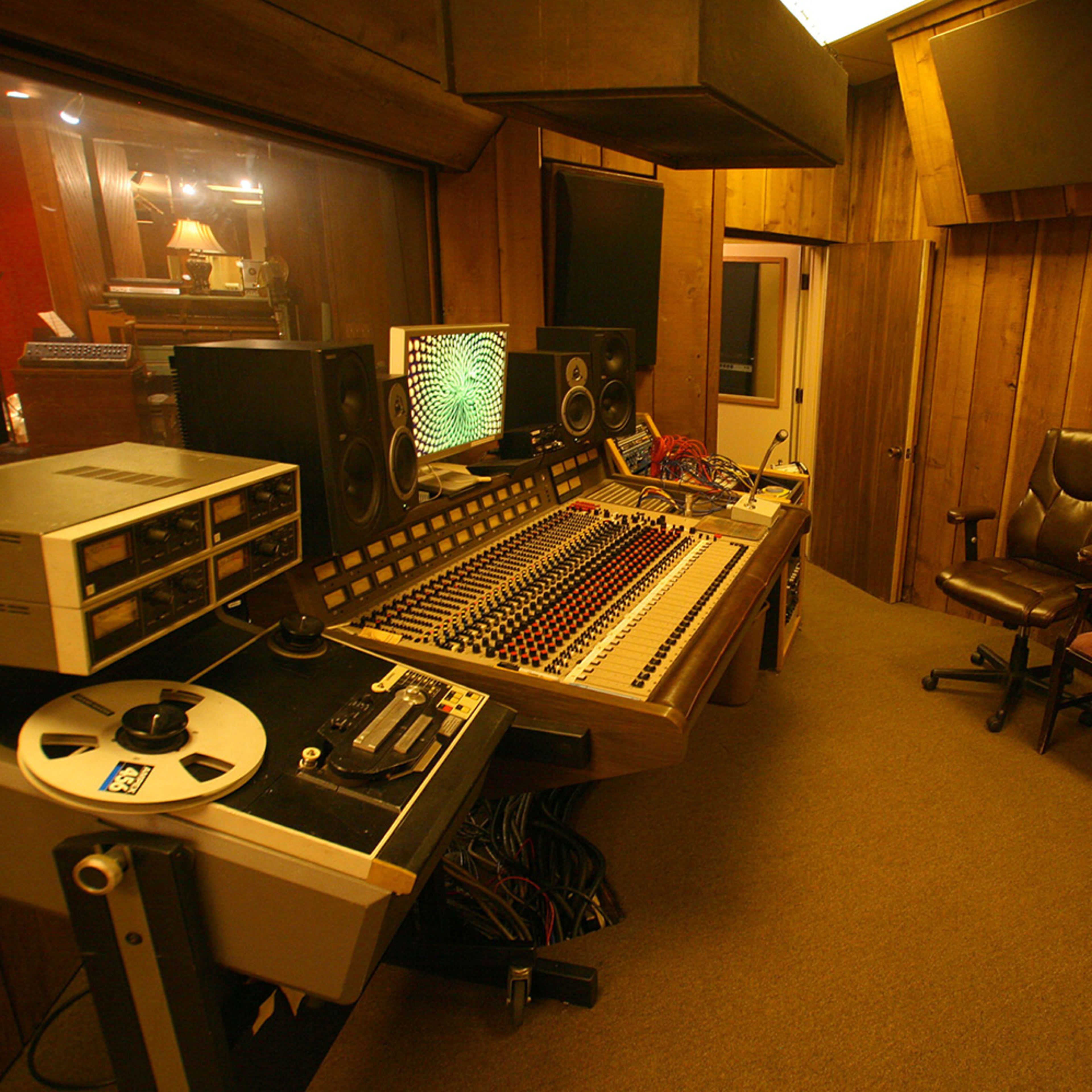 A recording studio with a mixing console, tape machine, and two chairs placed in a wooden-paneled room.