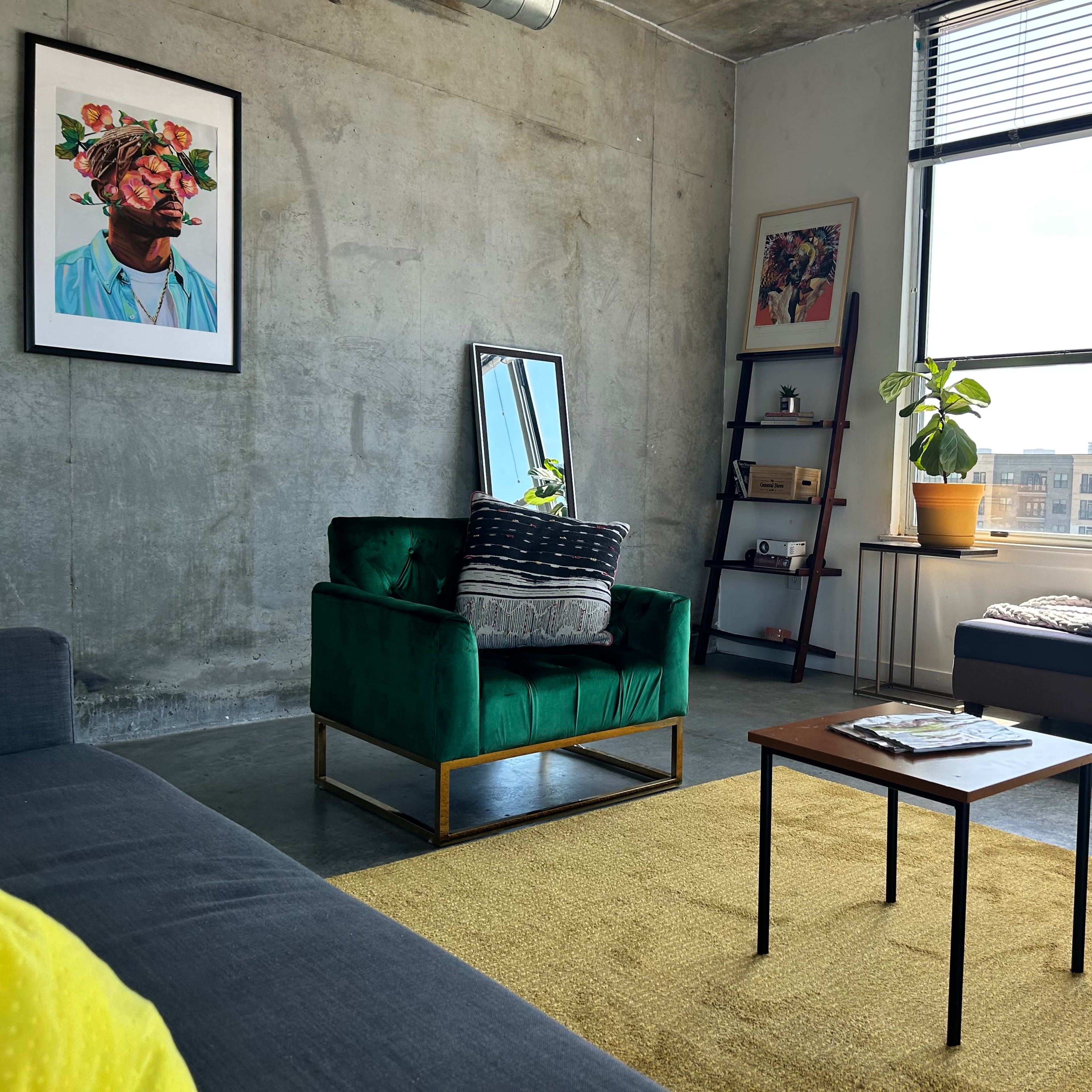 A modern living room featuring a green velvet chair, a wooden coffee table, a large window with natural light, and a mix of artwork on the walls.