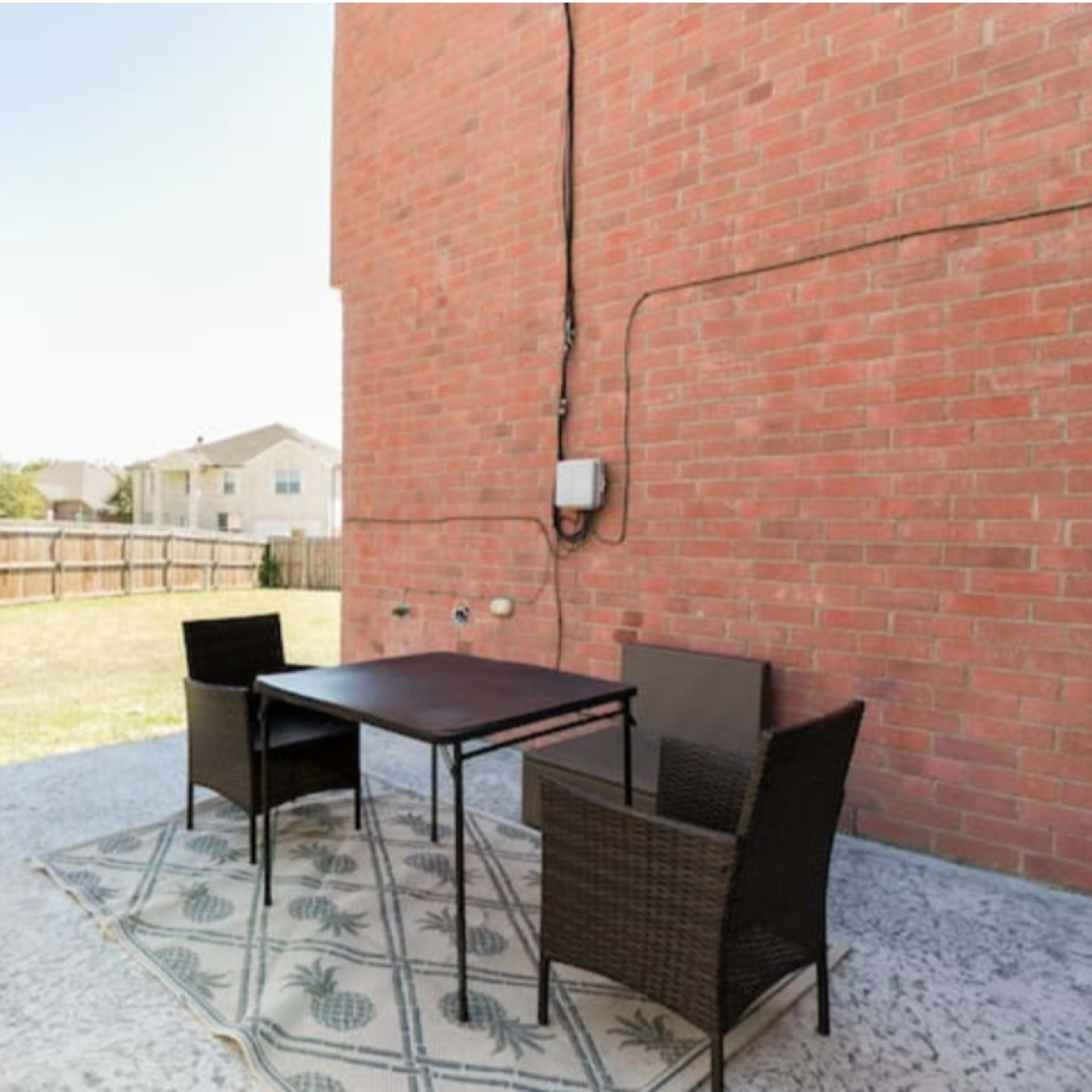A patio area features a black table and four chairs on a patterned rug, next to a grill and overlooking a grassy yard.