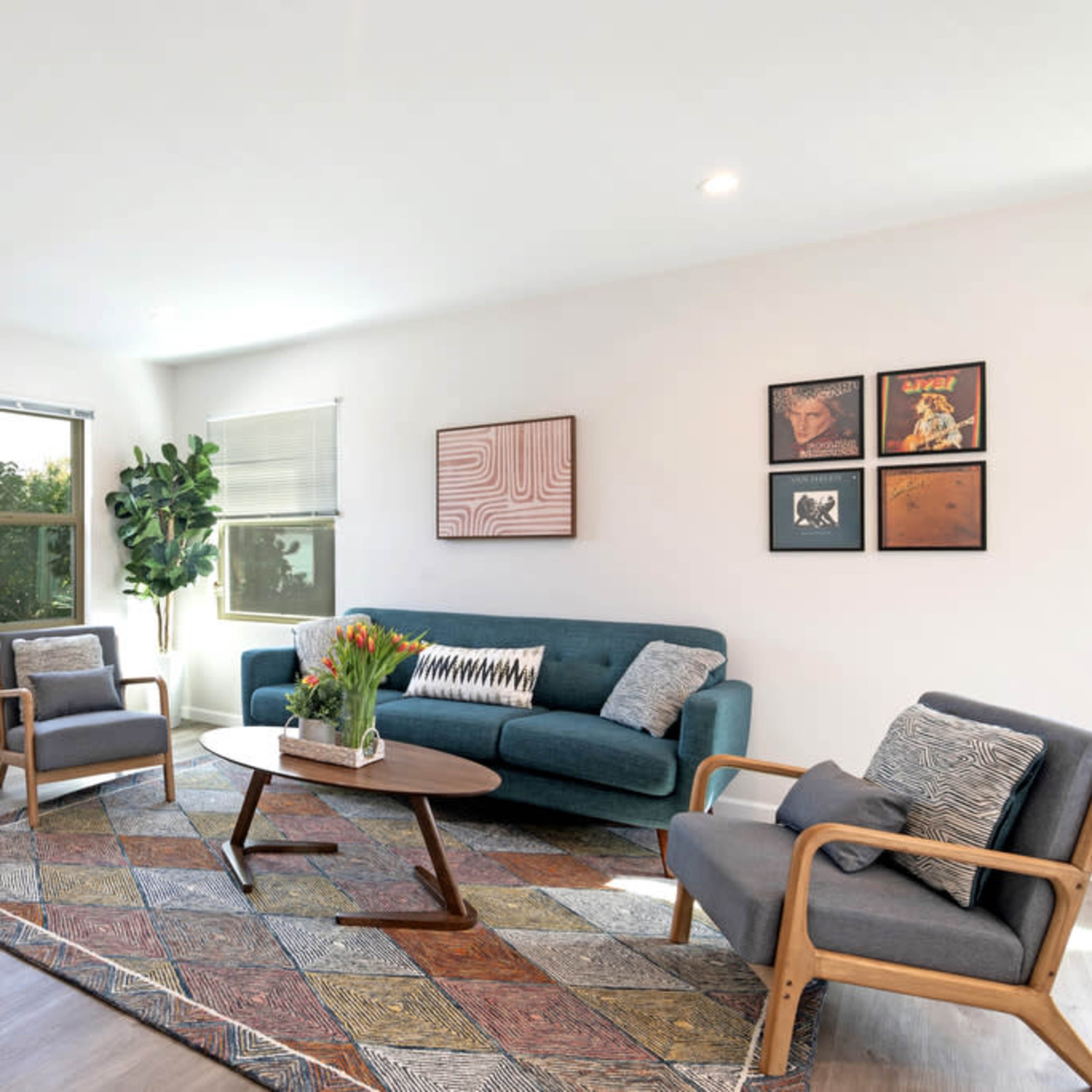 A modern living room features a green sofa, two wooden armchairs, a round coffee table, and a colorful patterned rug, with large windows allowing natural light.