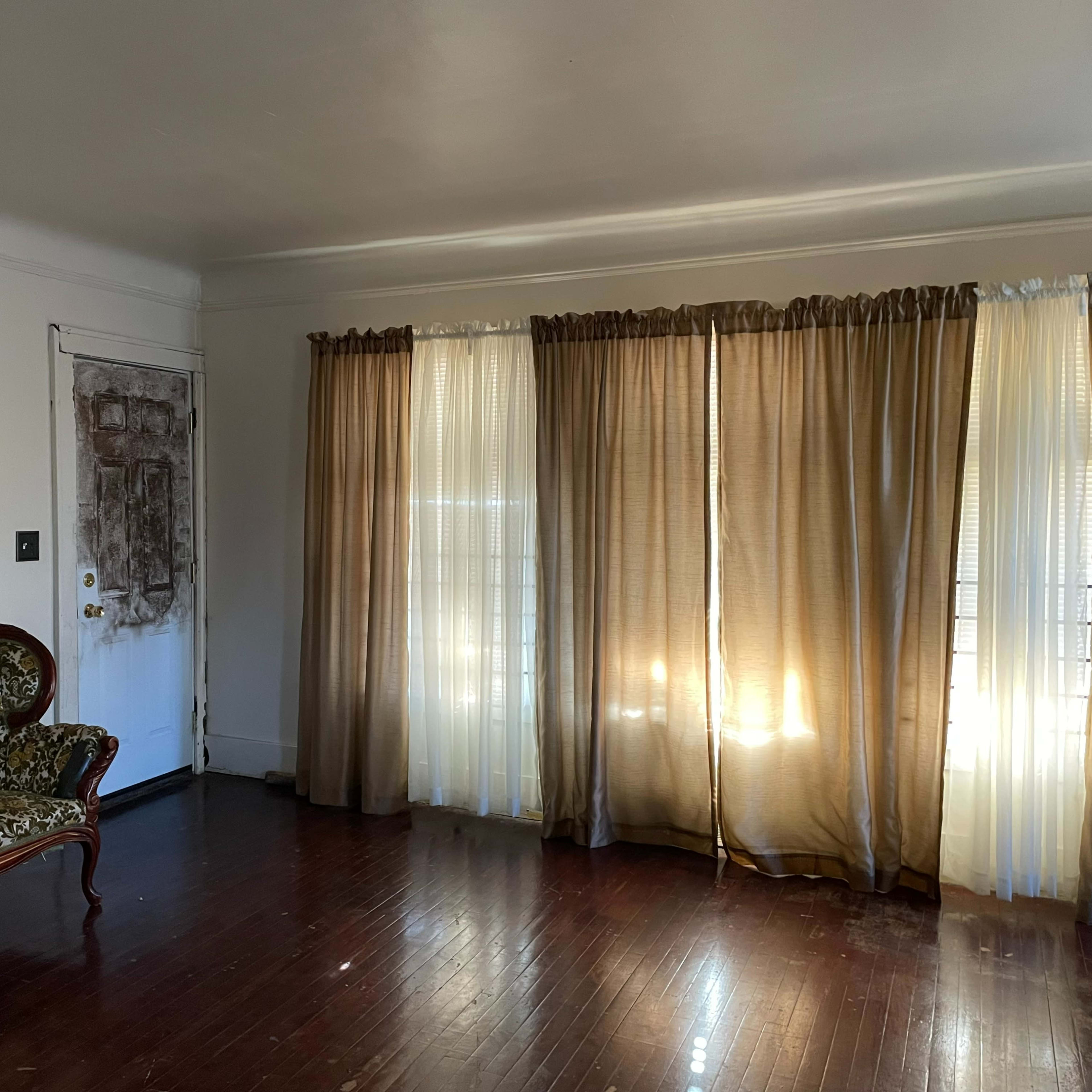 An empty room with a patterned couch, a weathered door, and light-colored curtains allowing sunlight to filter in.