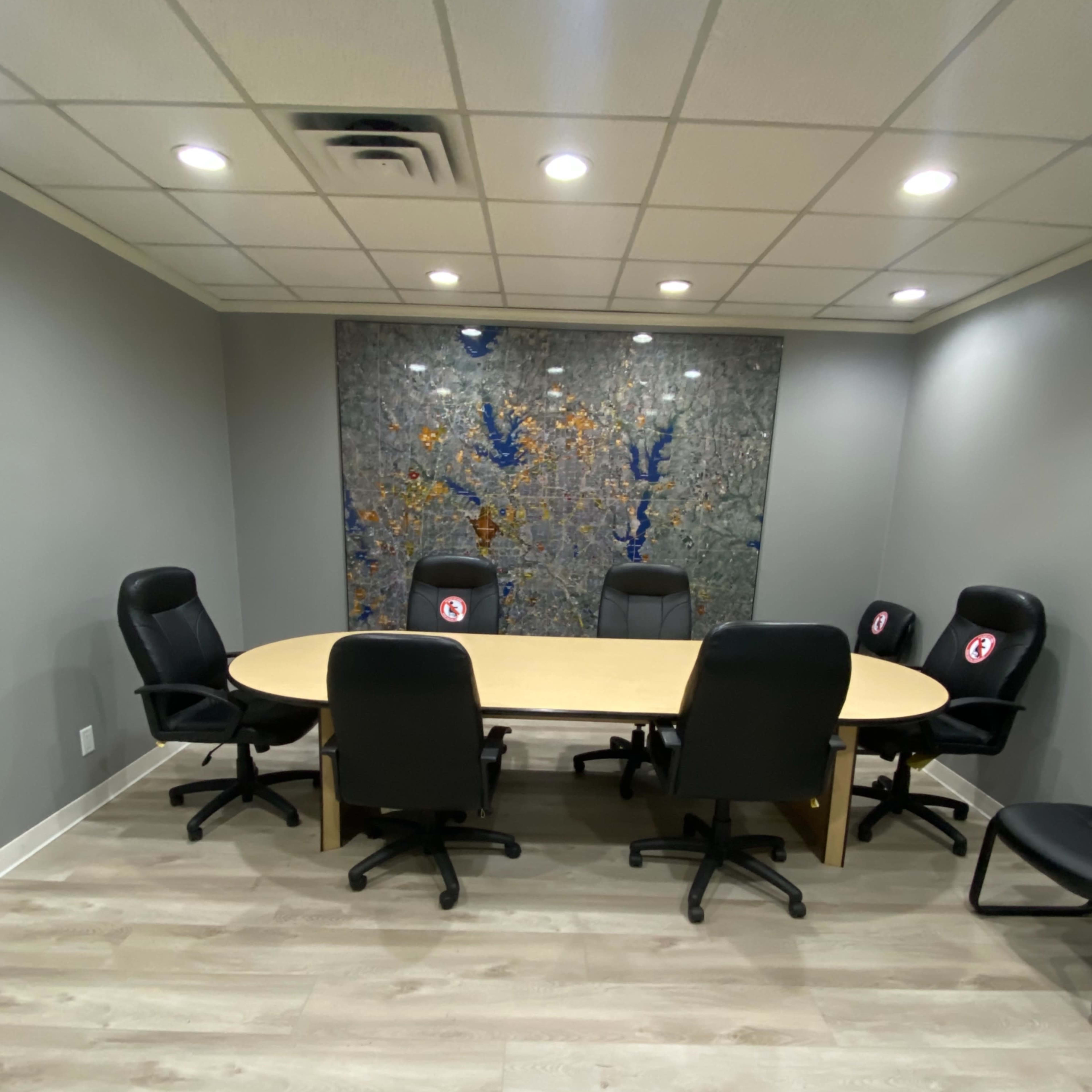 A conference room with a large oval table surrounded by black office chairs and a decorative wall in the background.