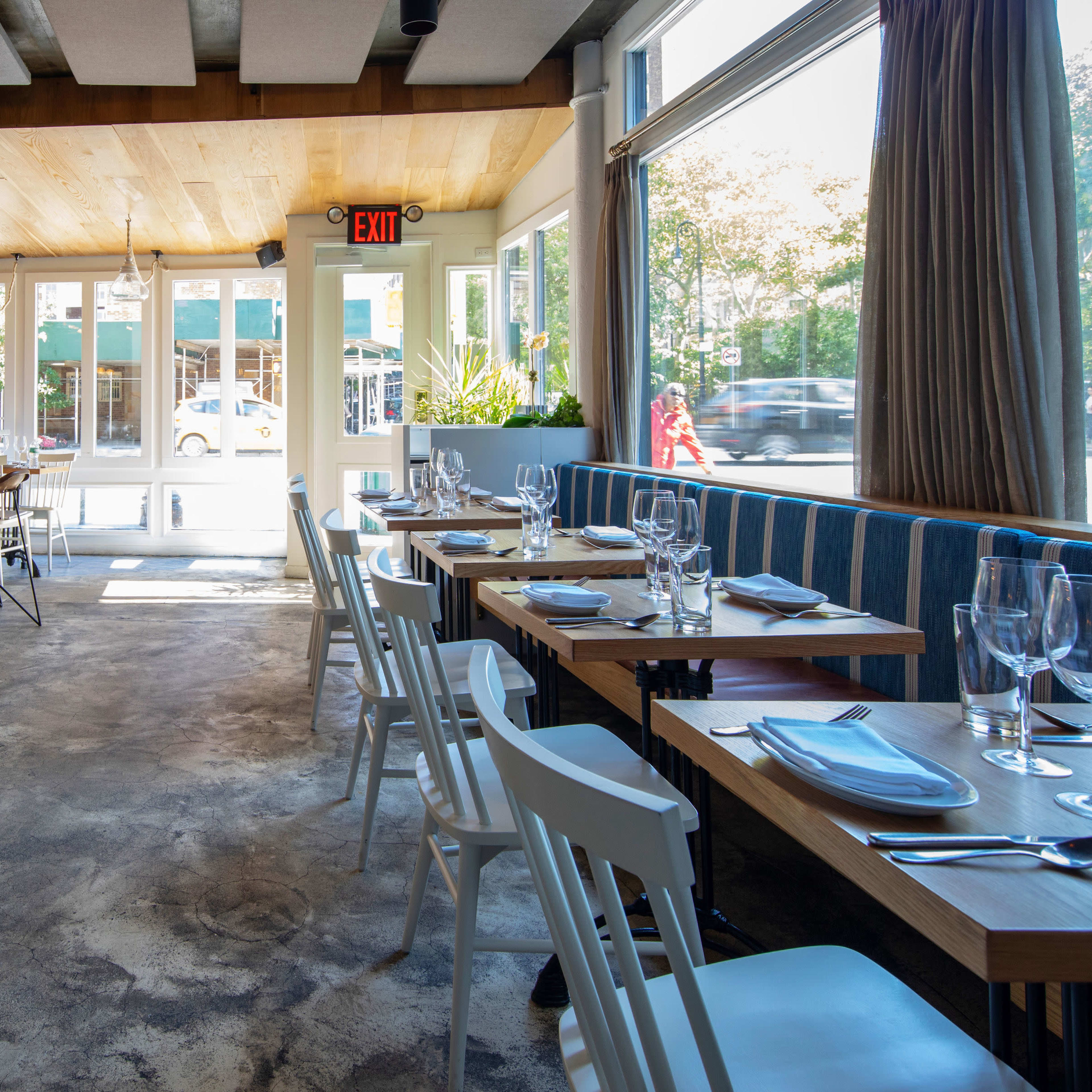 A modern restaurant interior with a bar on one side, wooden tables and chairs, and large windows letting in natural light.