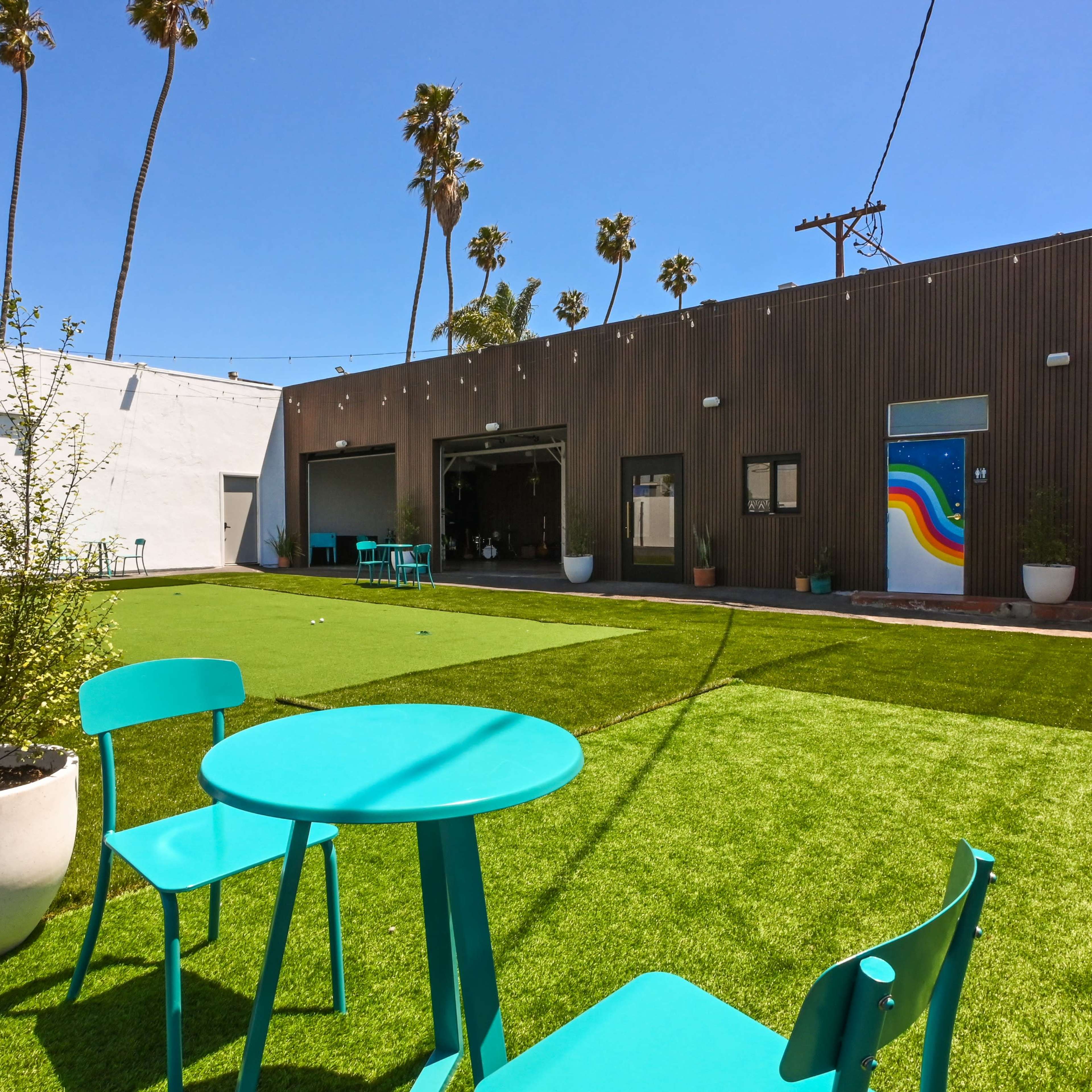A spacious outdoor area featuring artificial grass, two turquoise chairs, and buildings with colorful murals and palm trees in the background.