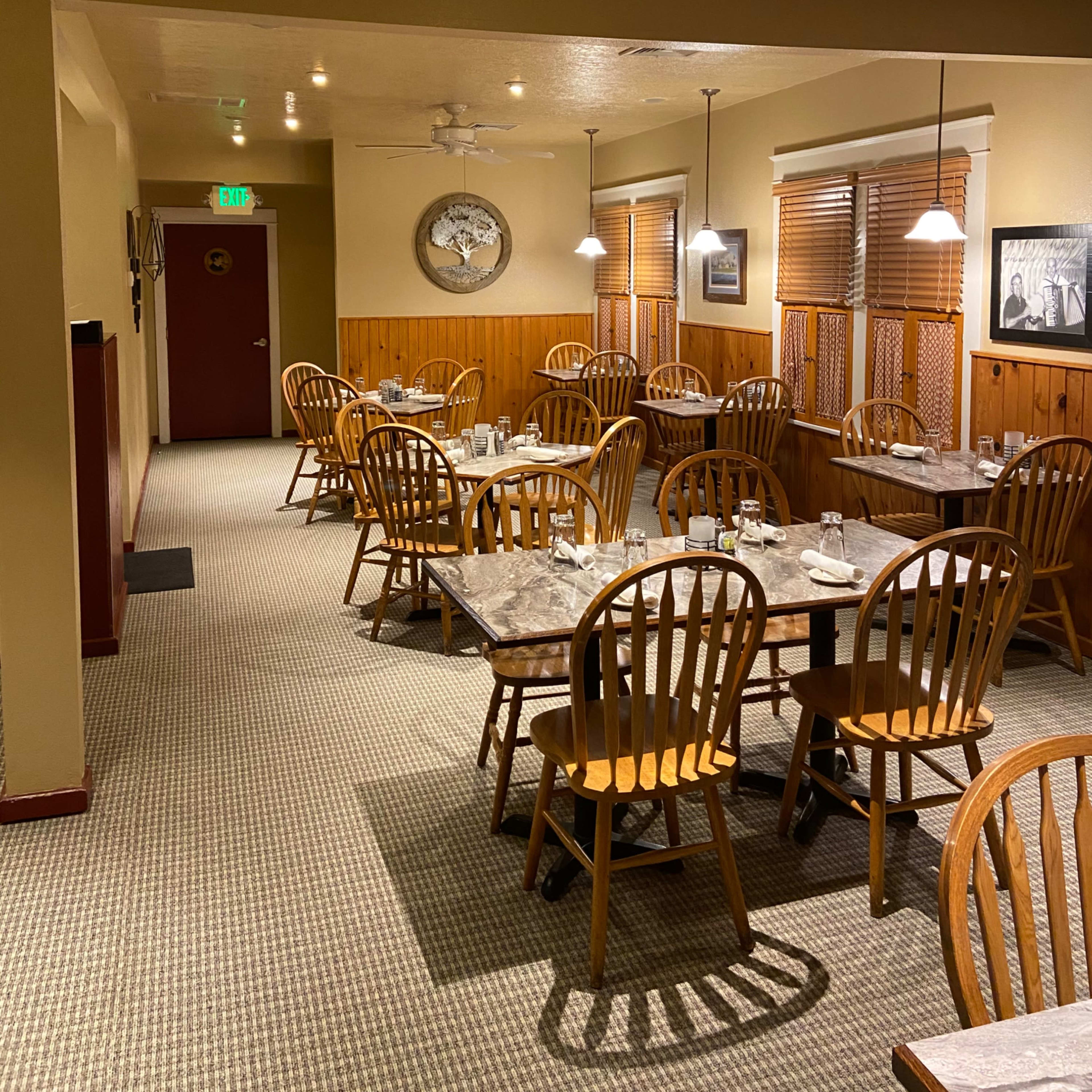 A quiet restaurant interior with wooden tables and chairs arranged in rows, featuring ceiling fans and large windows covered with blinds.