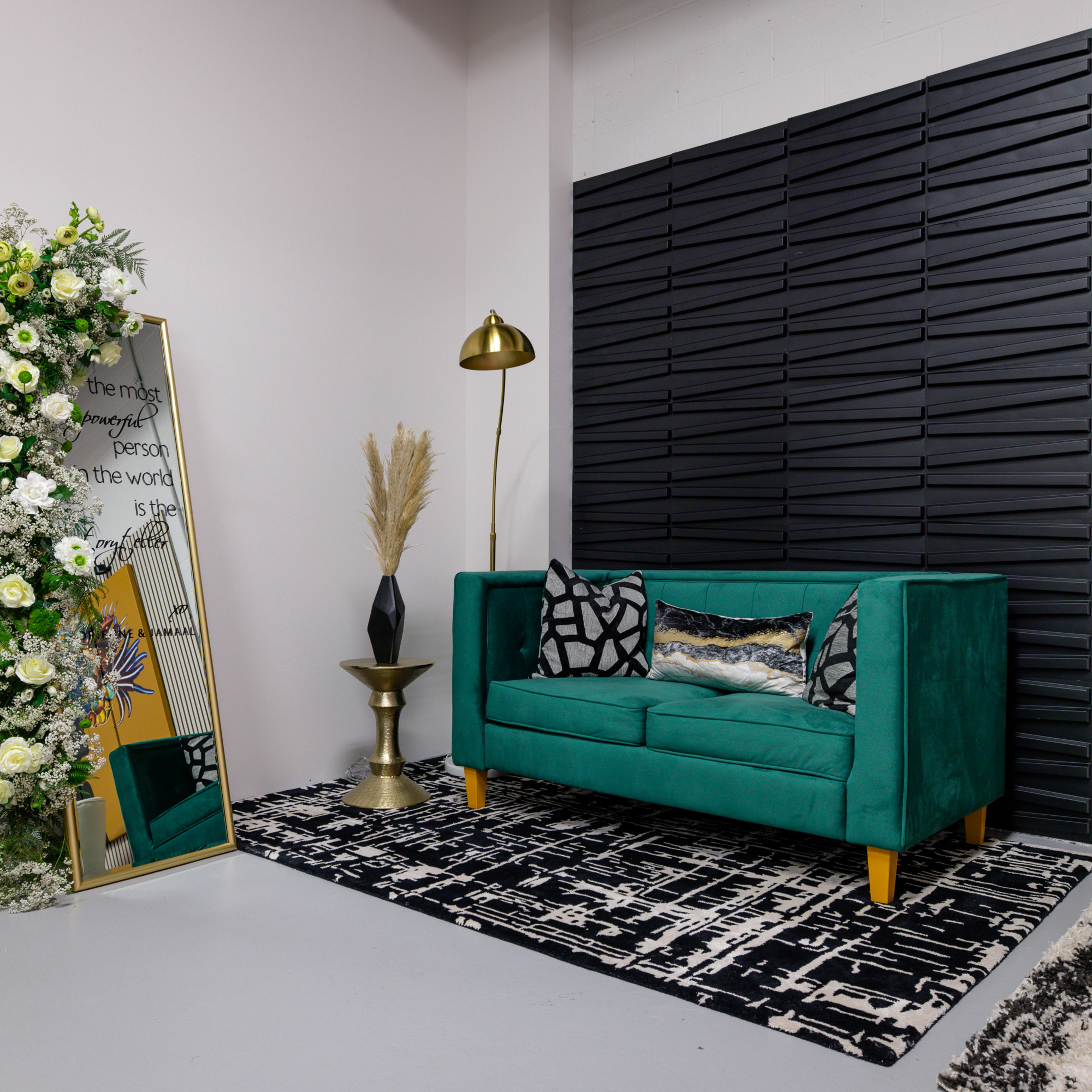 A green sofa with patterned cushions, a decorative mirror surrounded by flowers, a gold floor lamp, a potted plant, and a black-and-white patterned rug in a modern indoor setting.