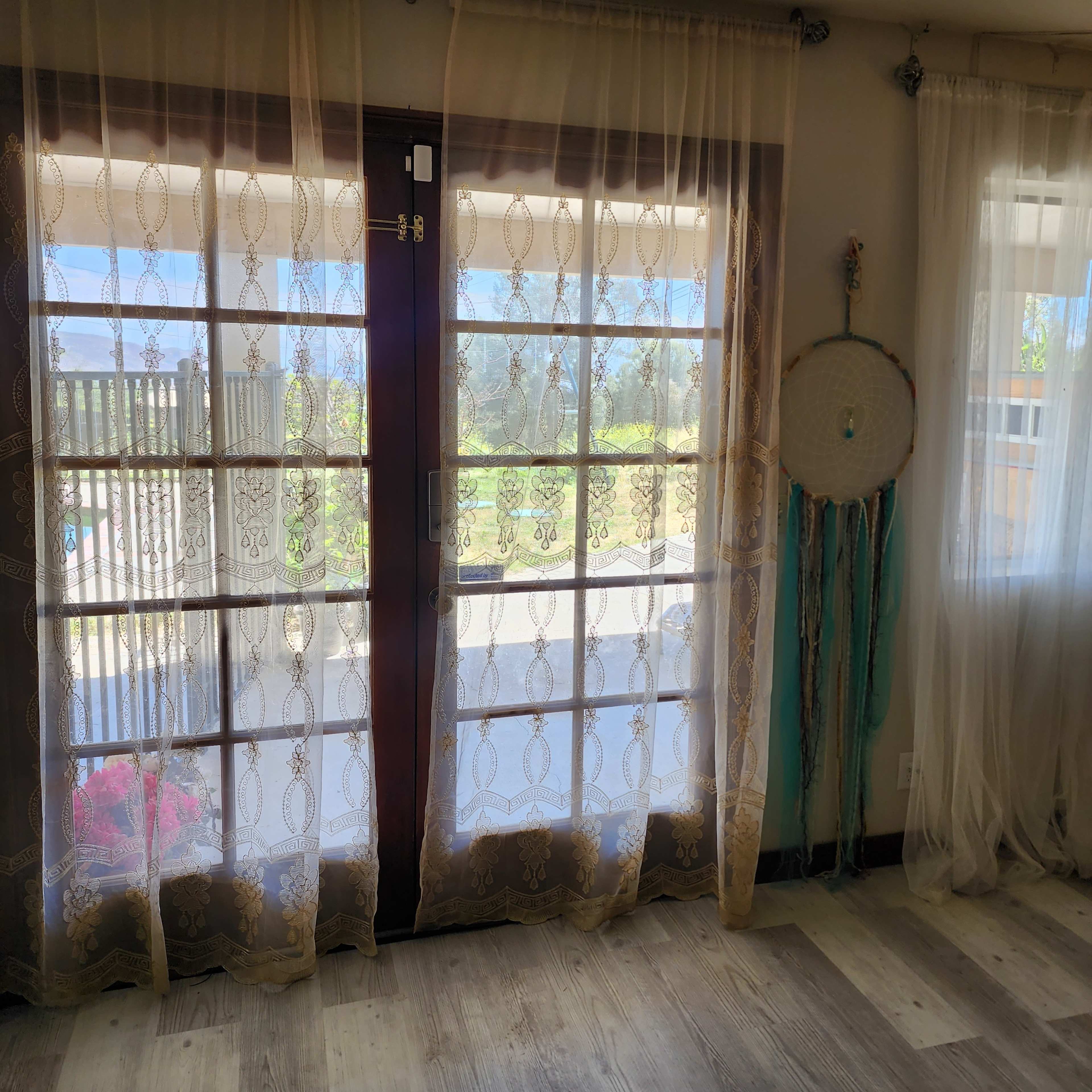 A room with sheer, patterned curtains over double glass doors leading to an outdoor view, and a dreamcatcher hanging nearby.