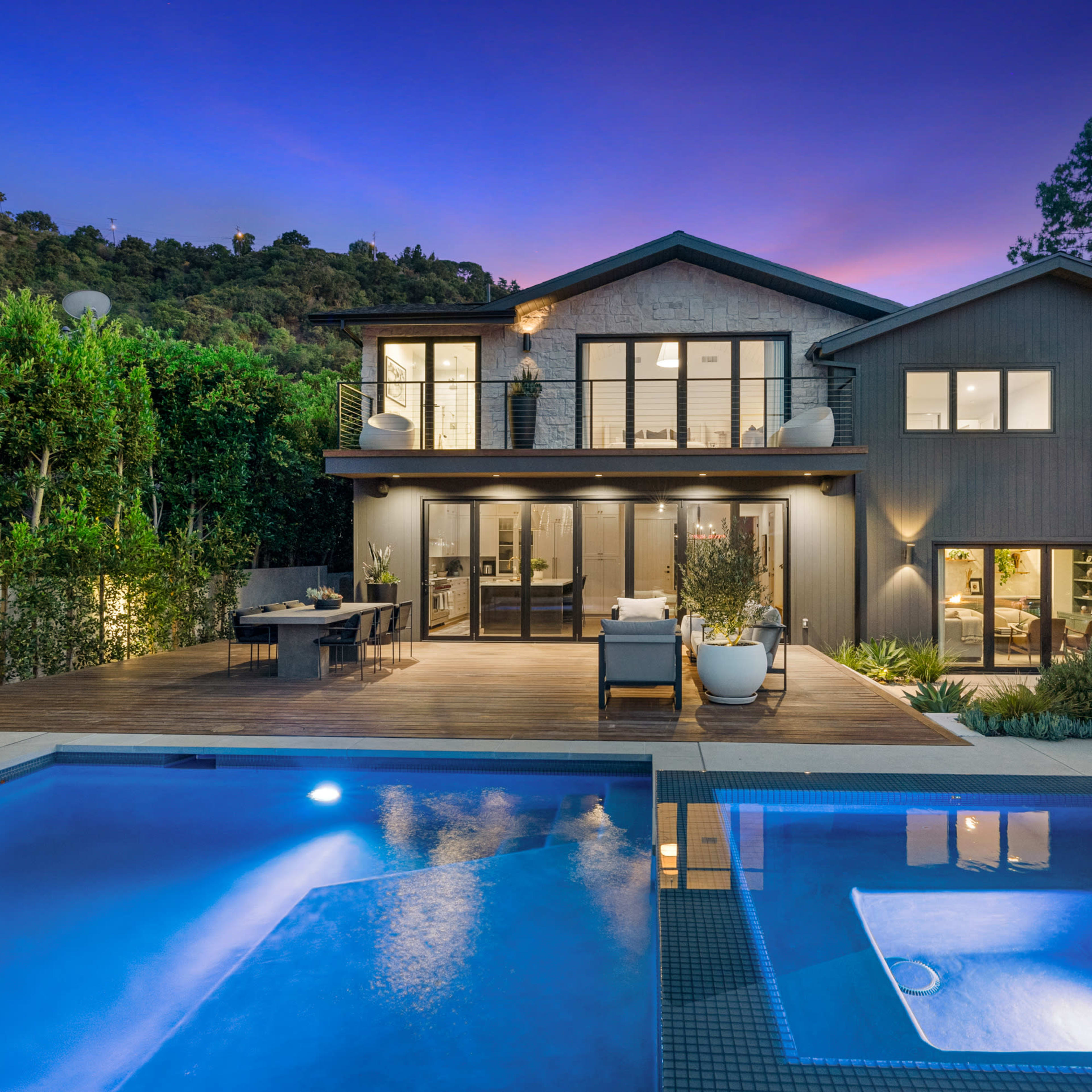 A modern two-story house with large glass doors overlooks a pool area surrounded by lush greenery and outdoor seating at twilight.
