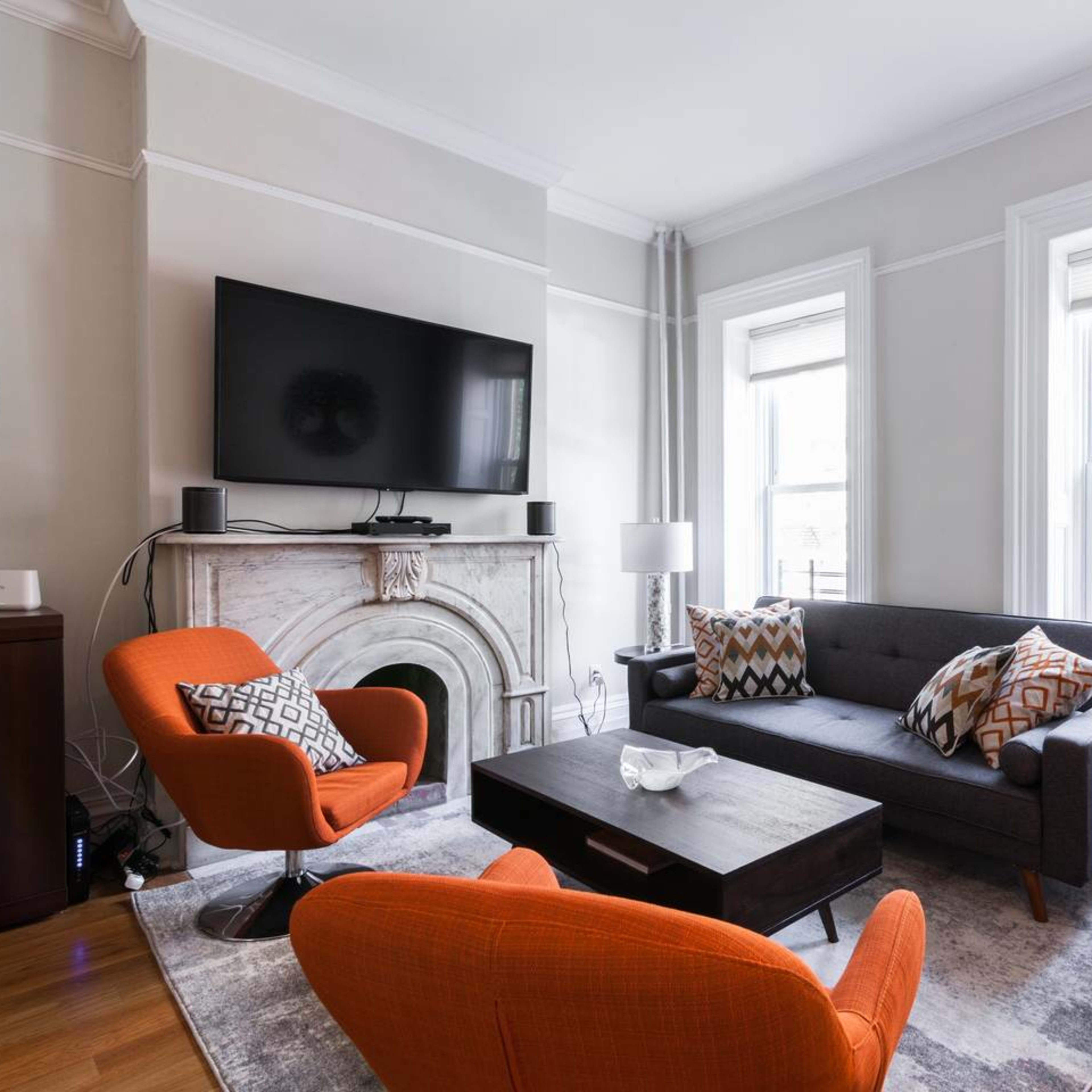 The living room features a gray couch, two orange armchairs, a dark wood coffee table, and a marble fireplace, with a television mounted on the wall above it.
