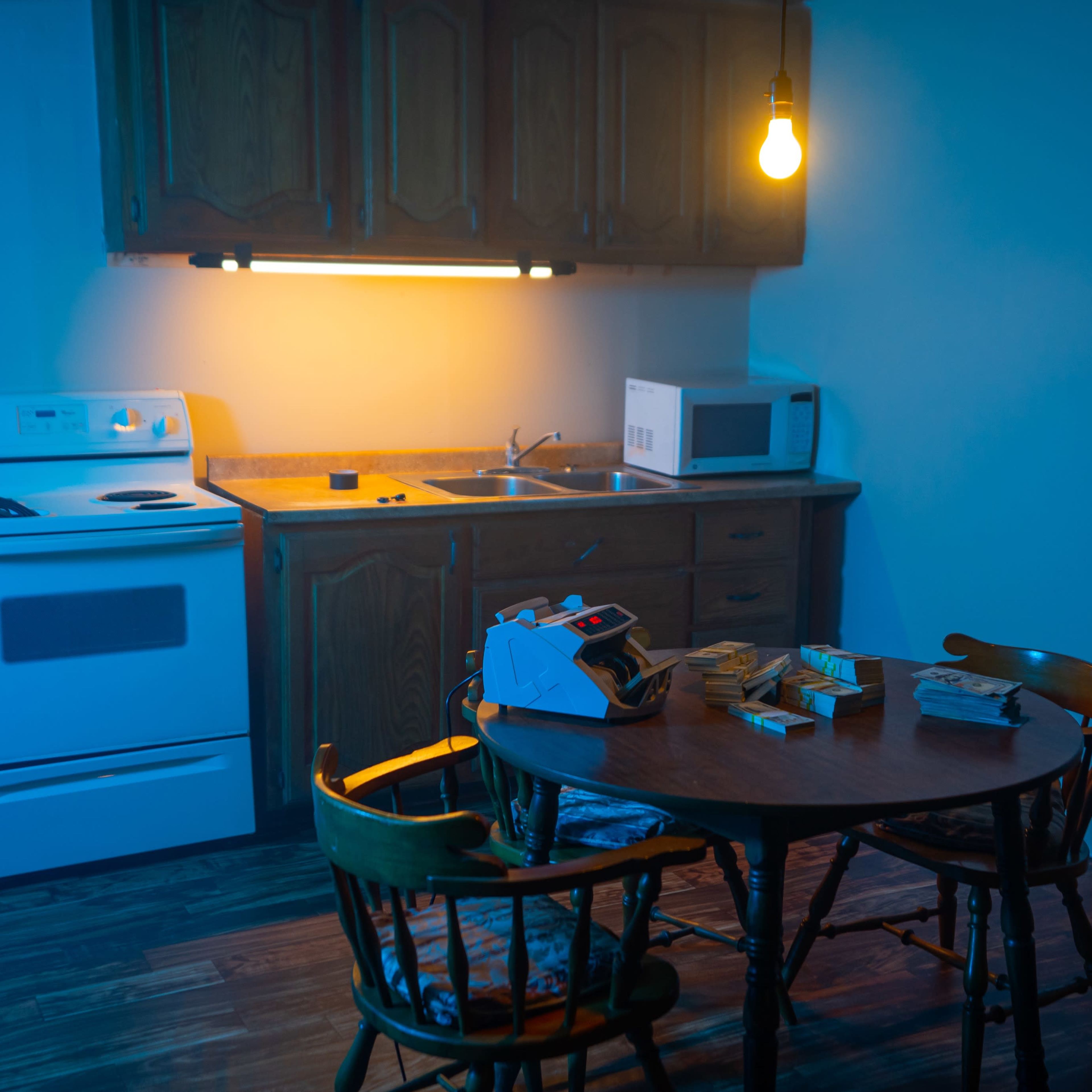 A dimly lit kitchen features a table with chairs, a stove, a microwave, and stacks of cash arranged on the table.