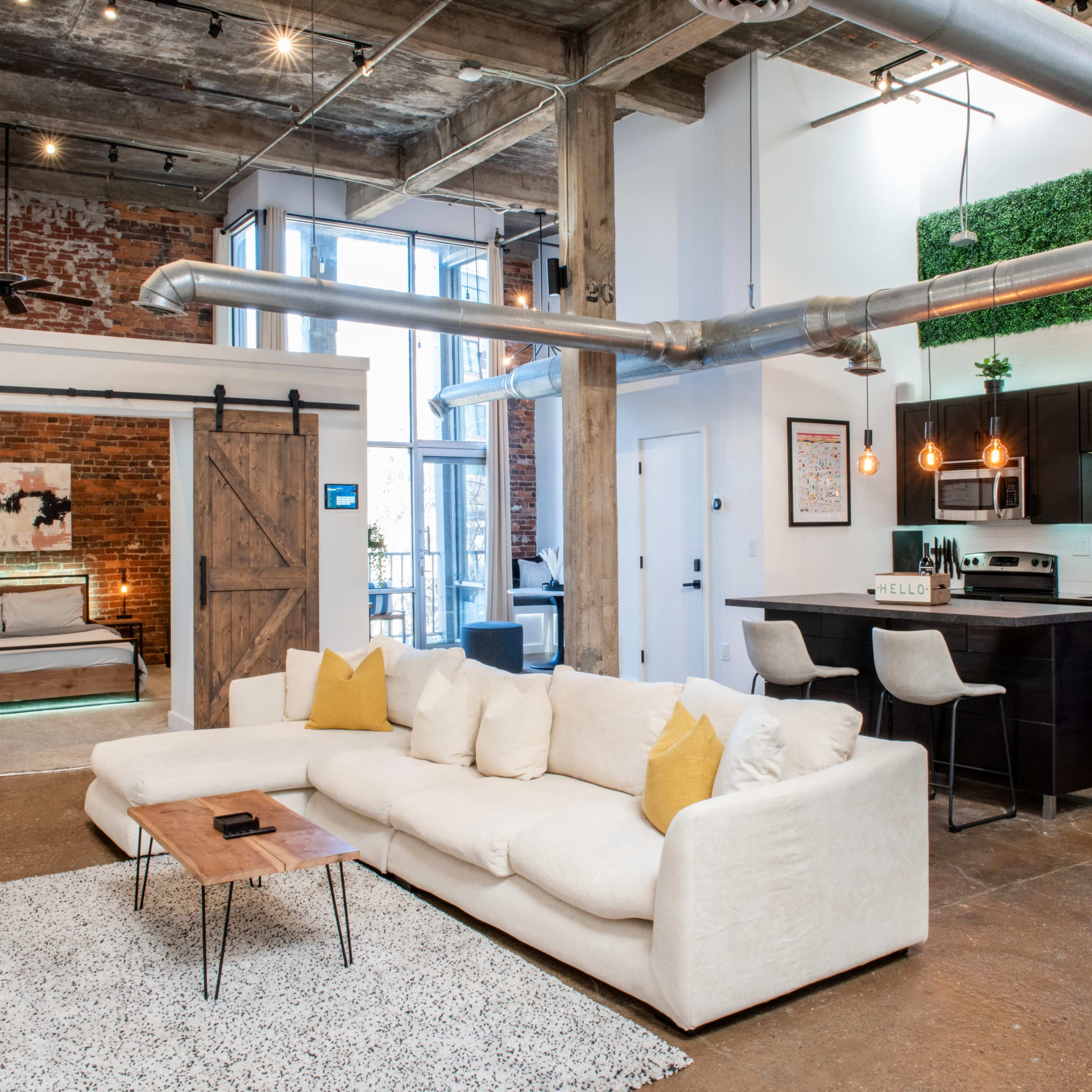 A modern open-concept living space with a white sectional sofa, a wooden coffee table, and a kitchen area featuring dark cabinetry and industrial elements.