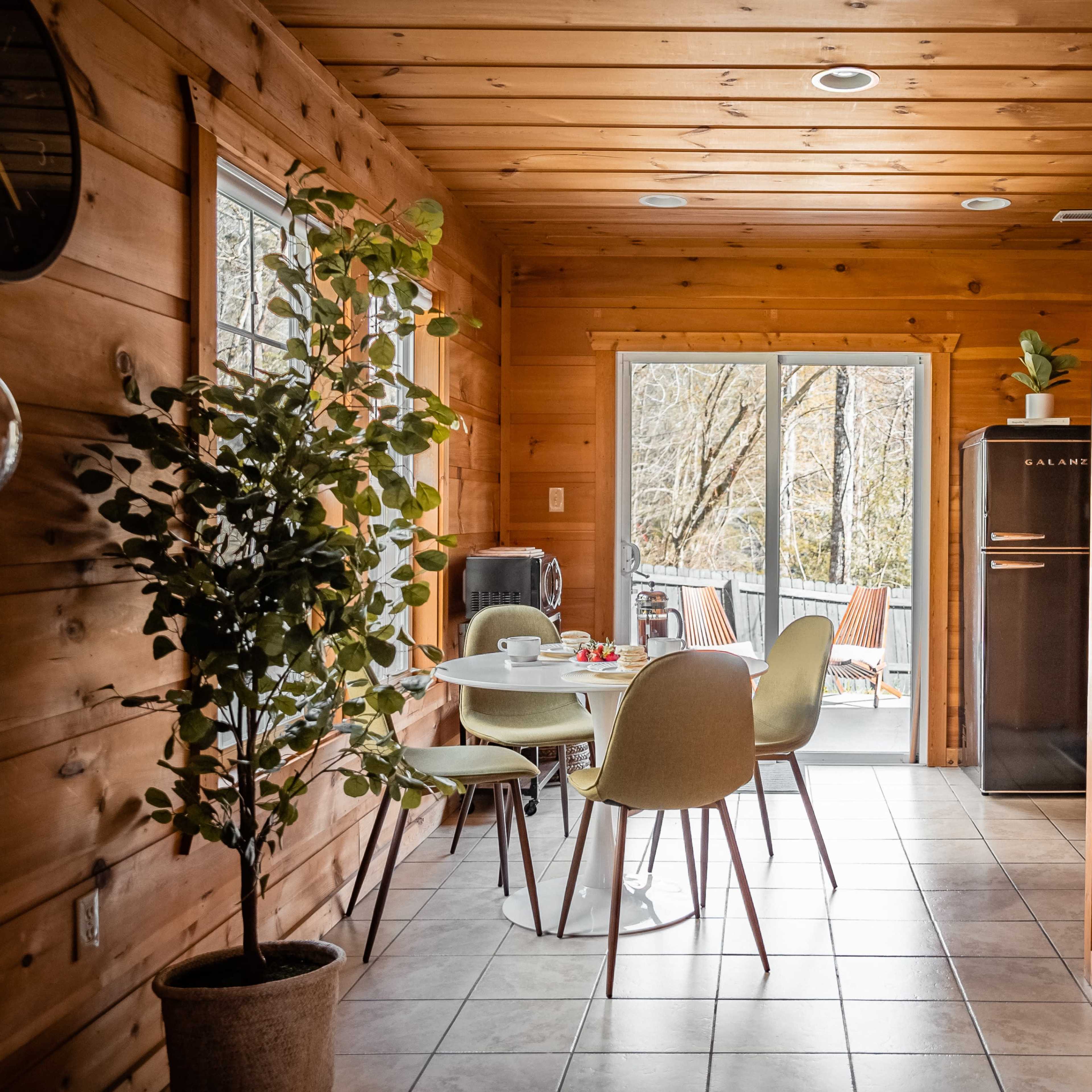 A dining area with a small round table and chairs is situated in a wooden-paneled room, leading to a door that opens to an outdoor seating area.