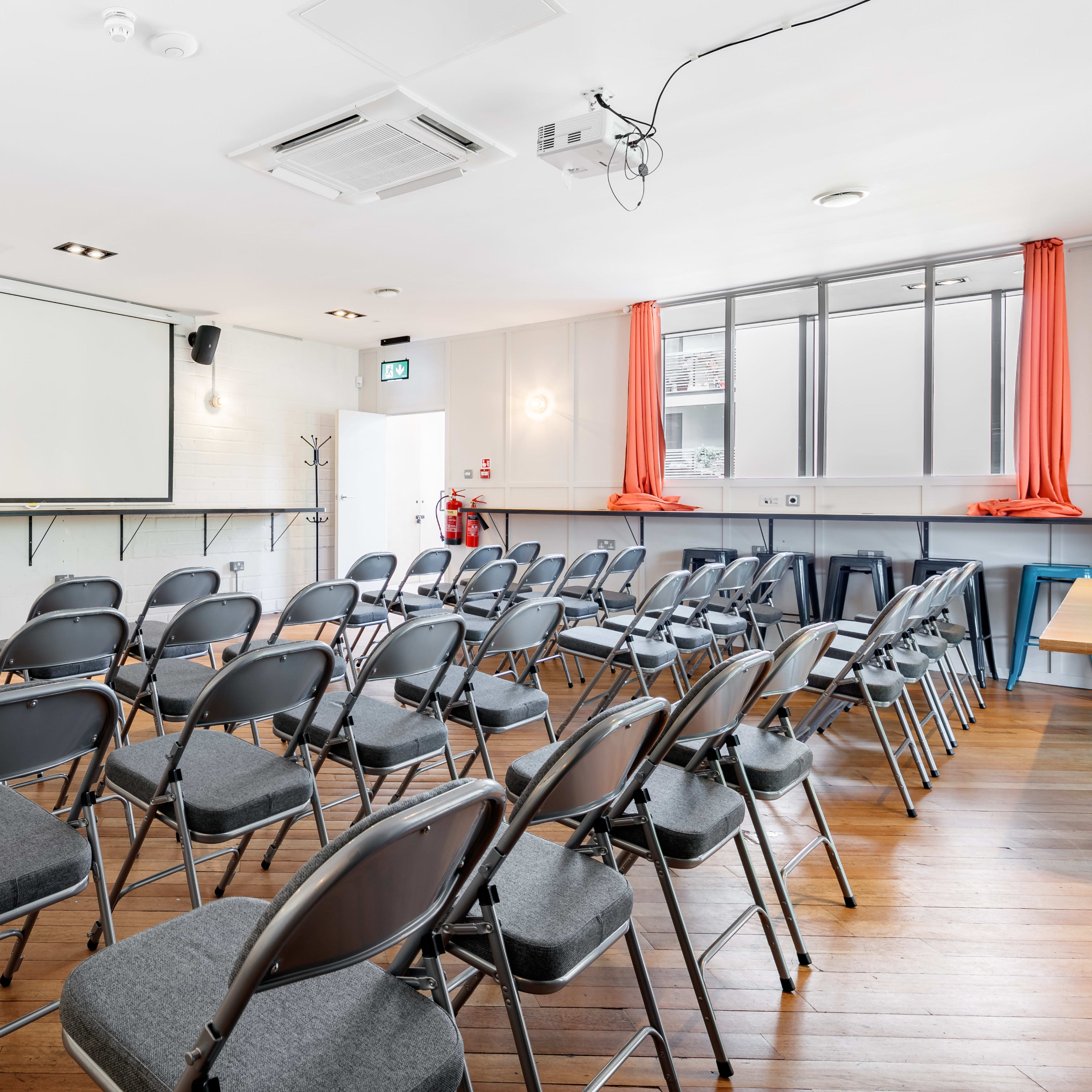 A meeting room with rows of gray chairs facing a blank projector screen, flanked by orange curtains and wooden flooring.