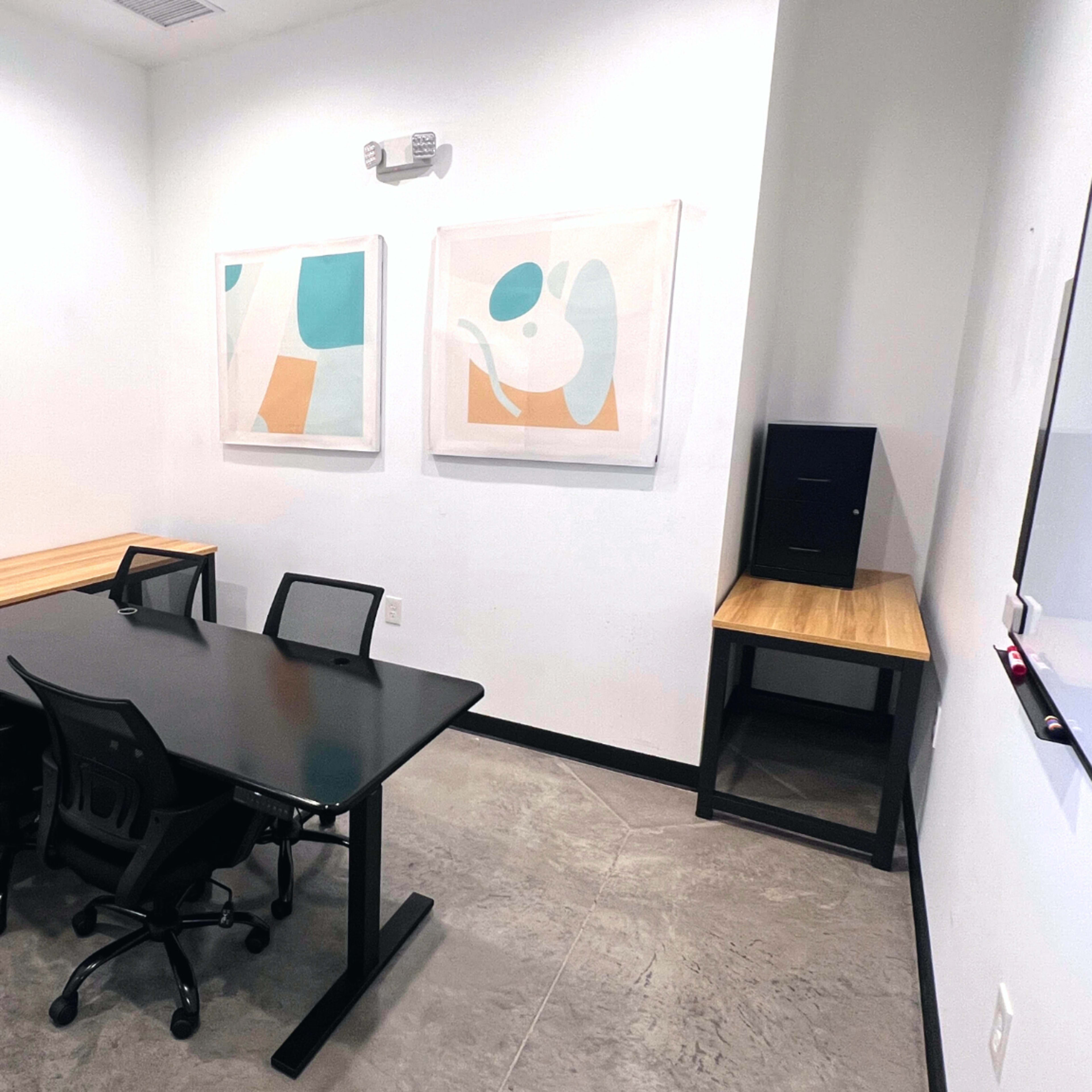 A small conference room with a black table and four chairs, two wall art pieces, and a side table against a white wall.