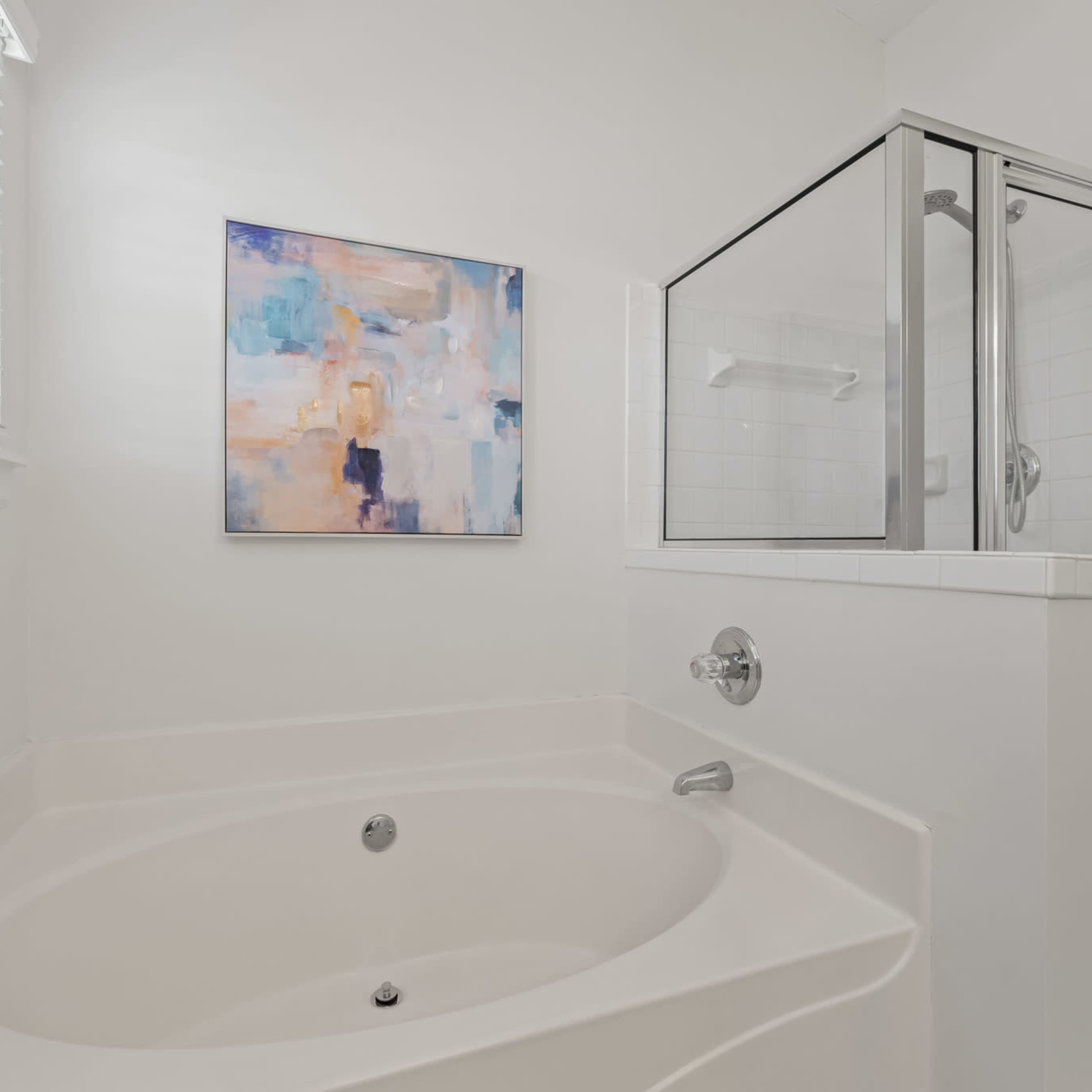 A bathroom features a white tub and a glass-enclosed shower, with light-colored walls and a piece of abstract artwork hanging above.