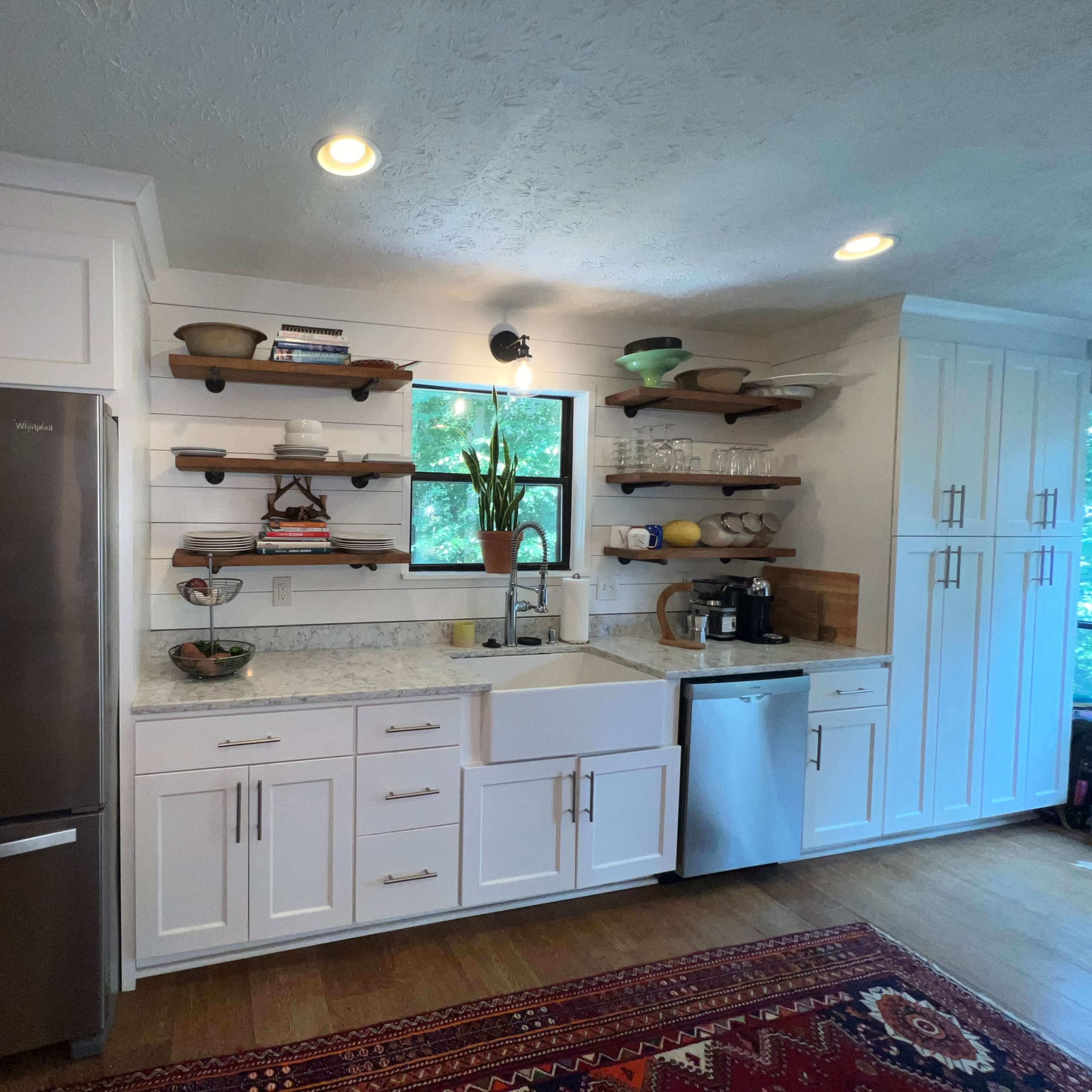 A modern kitchen with white cabinetry, a farmhouse sink, stainless steel appliances, and open shelves displaying dishware and decorative items.