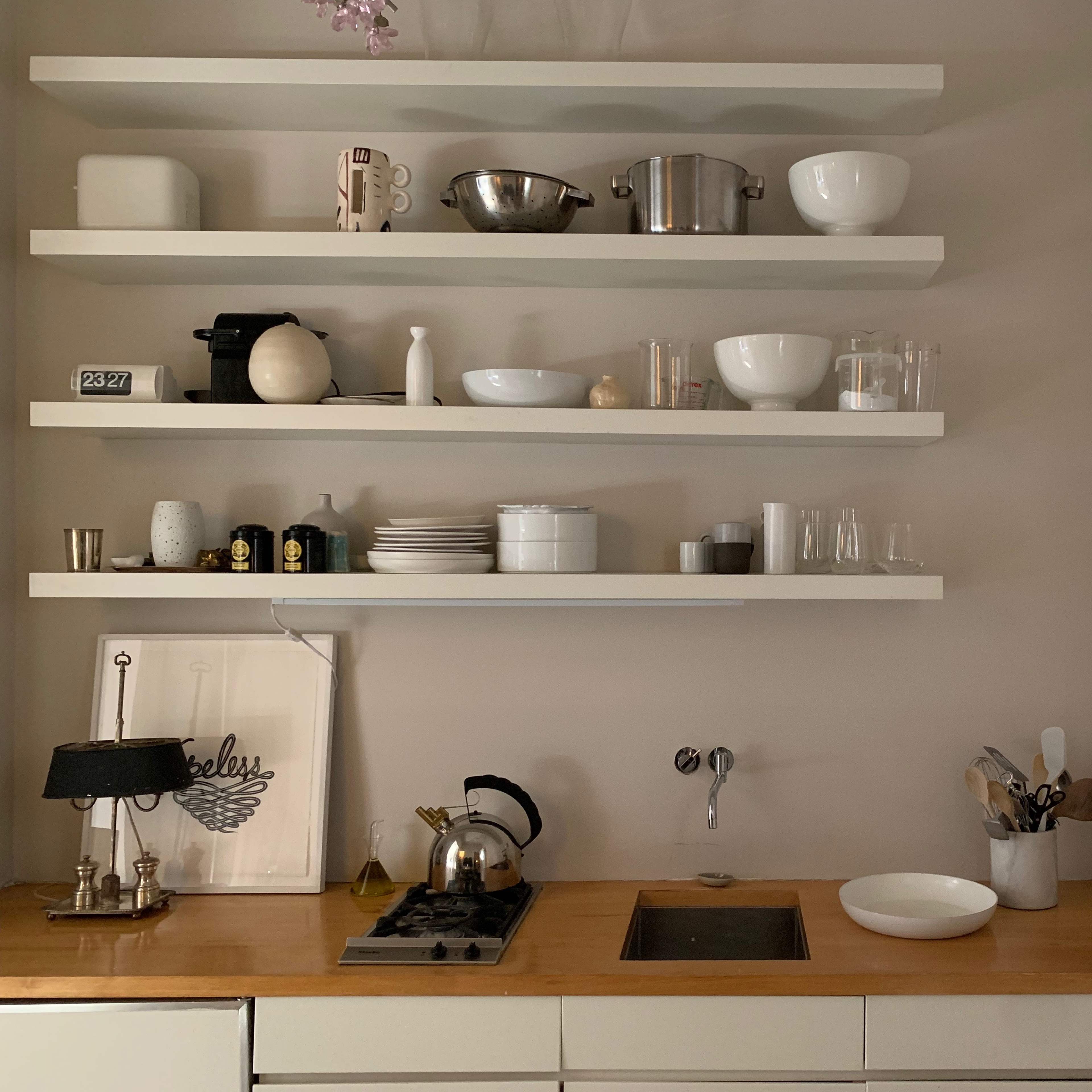 A minimalist kitchen with wooden countertops, white shelves displaying various kitchenware, and a stove with a kettle.
