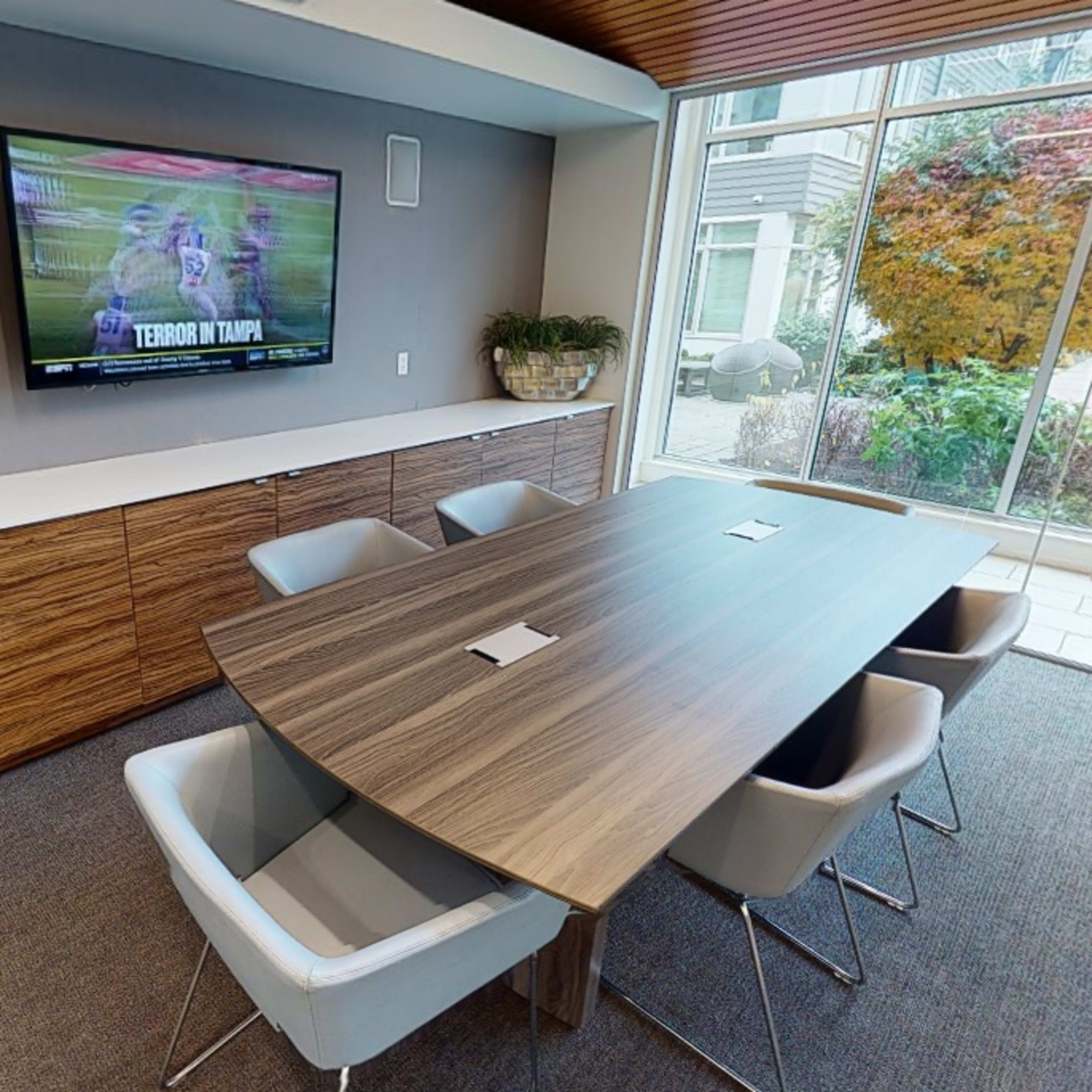 A modern conference room features a long wooden table surrounded by upholstered chairs and a television mounted on the wall displaying a sports broadcast.