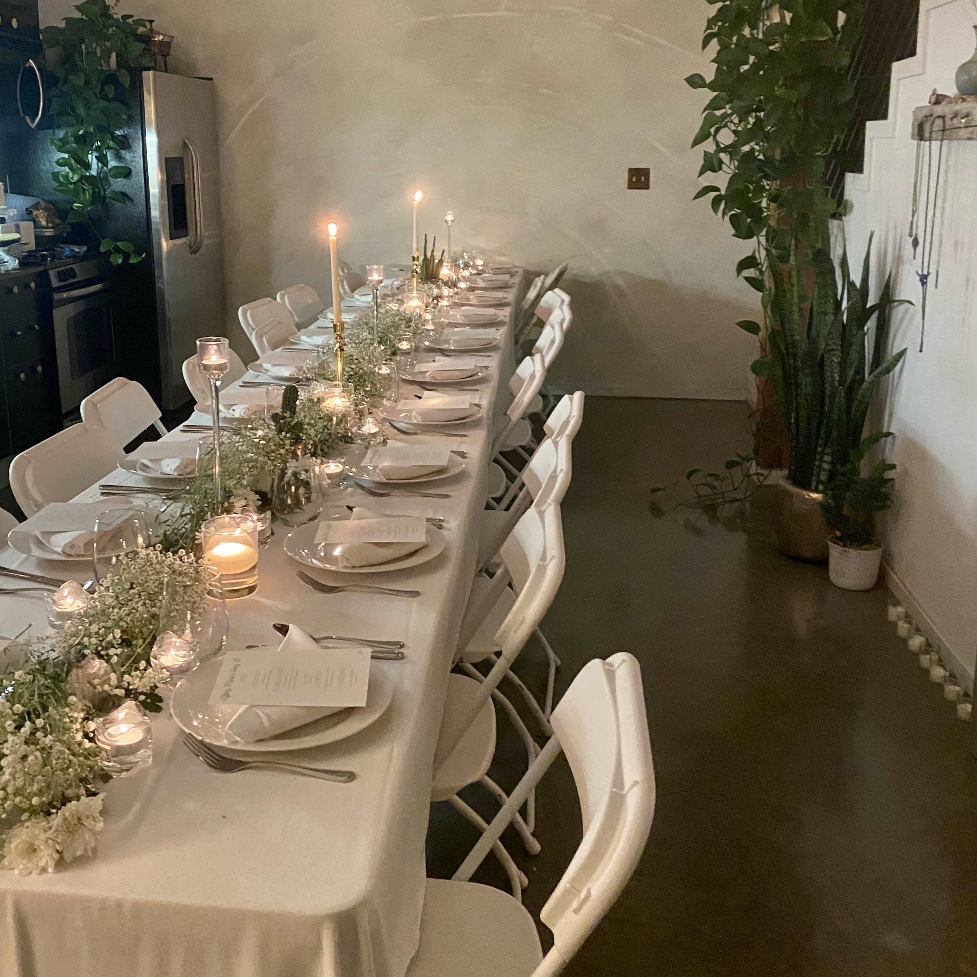 A long dining table is elegantly set with candles and flowers in a softly lit room featuring plants and a kitchen in the background.