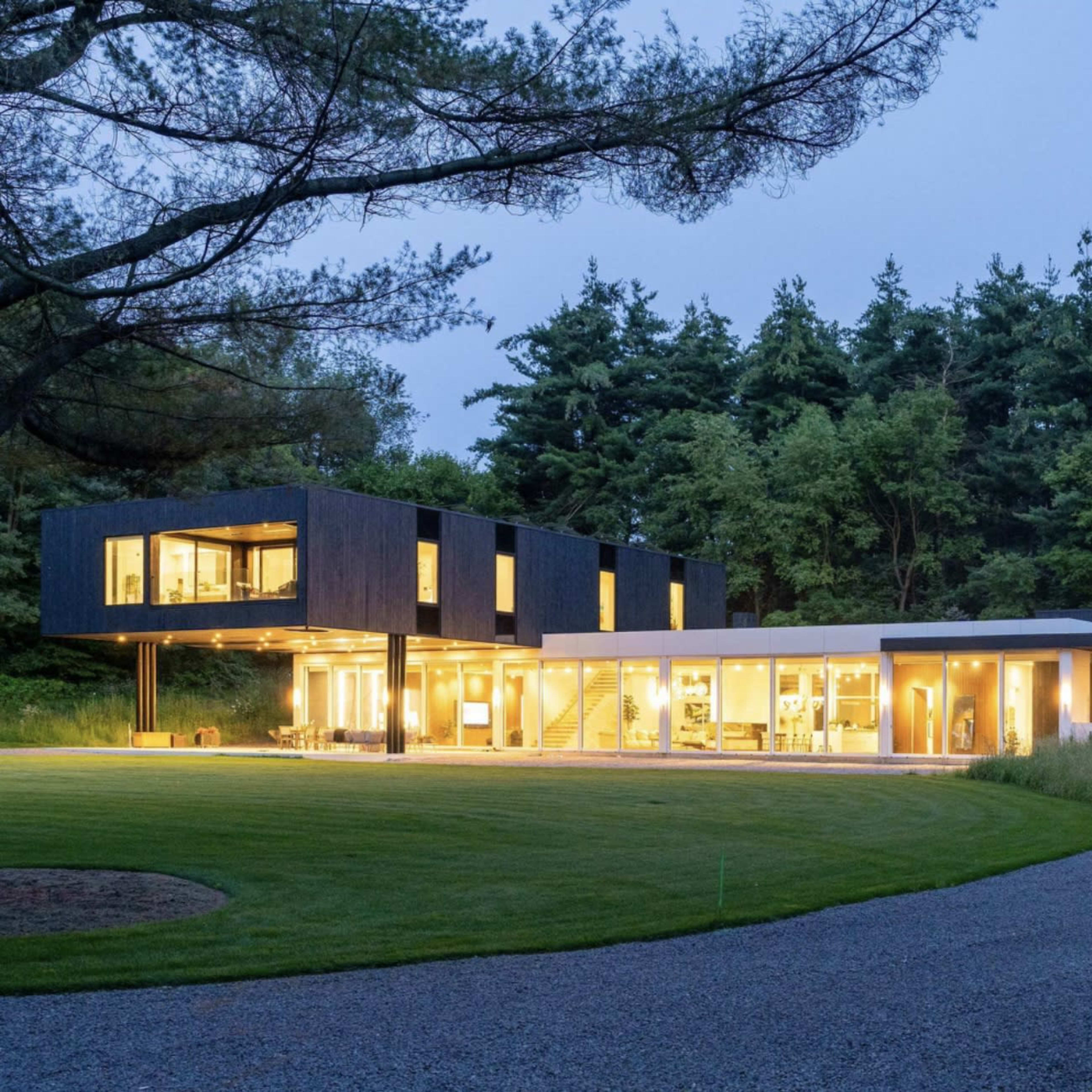 A modern two-story house with large windows, situated in a landscaped area with trees and a gravel driveway.