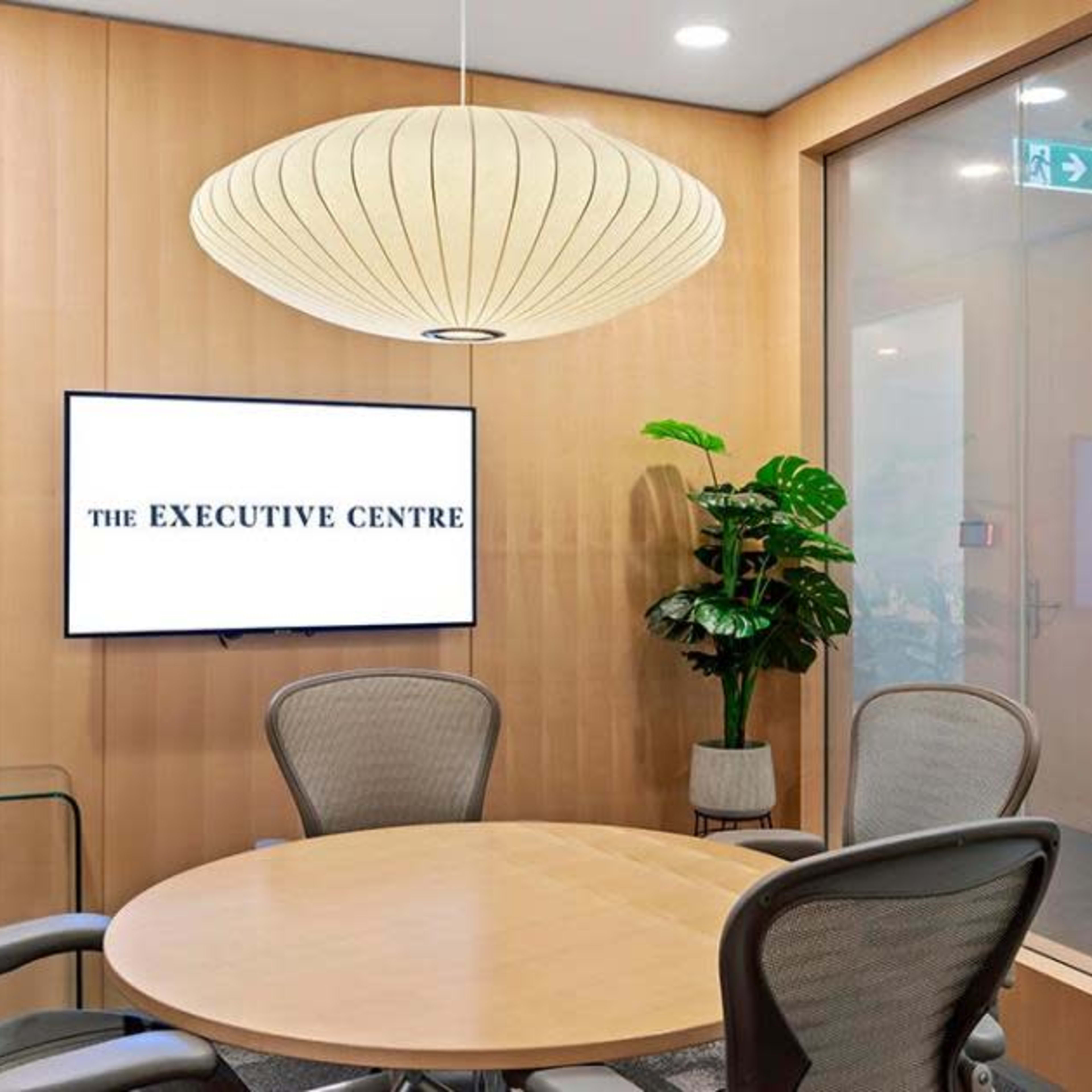 A minimalist meeting room features a round wooden table, ergonomic chairs, a wall-mounted television displaying "THE EXECUTIVE CENTRE," and a large pendant light above.