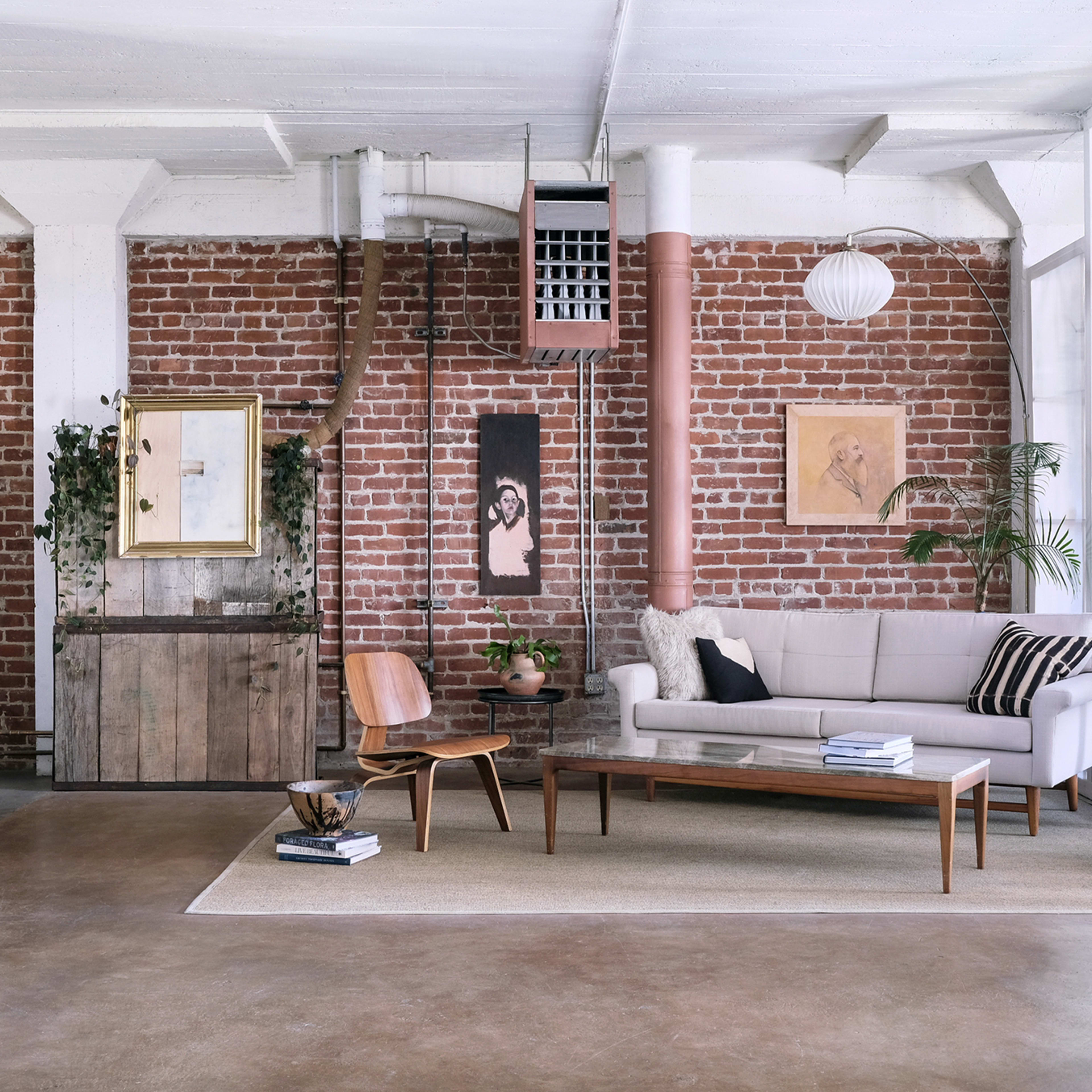 A minimalistic living room with a brick wall, a light-colored couch, a wooden coffee table, and various decorative plants and artwork.