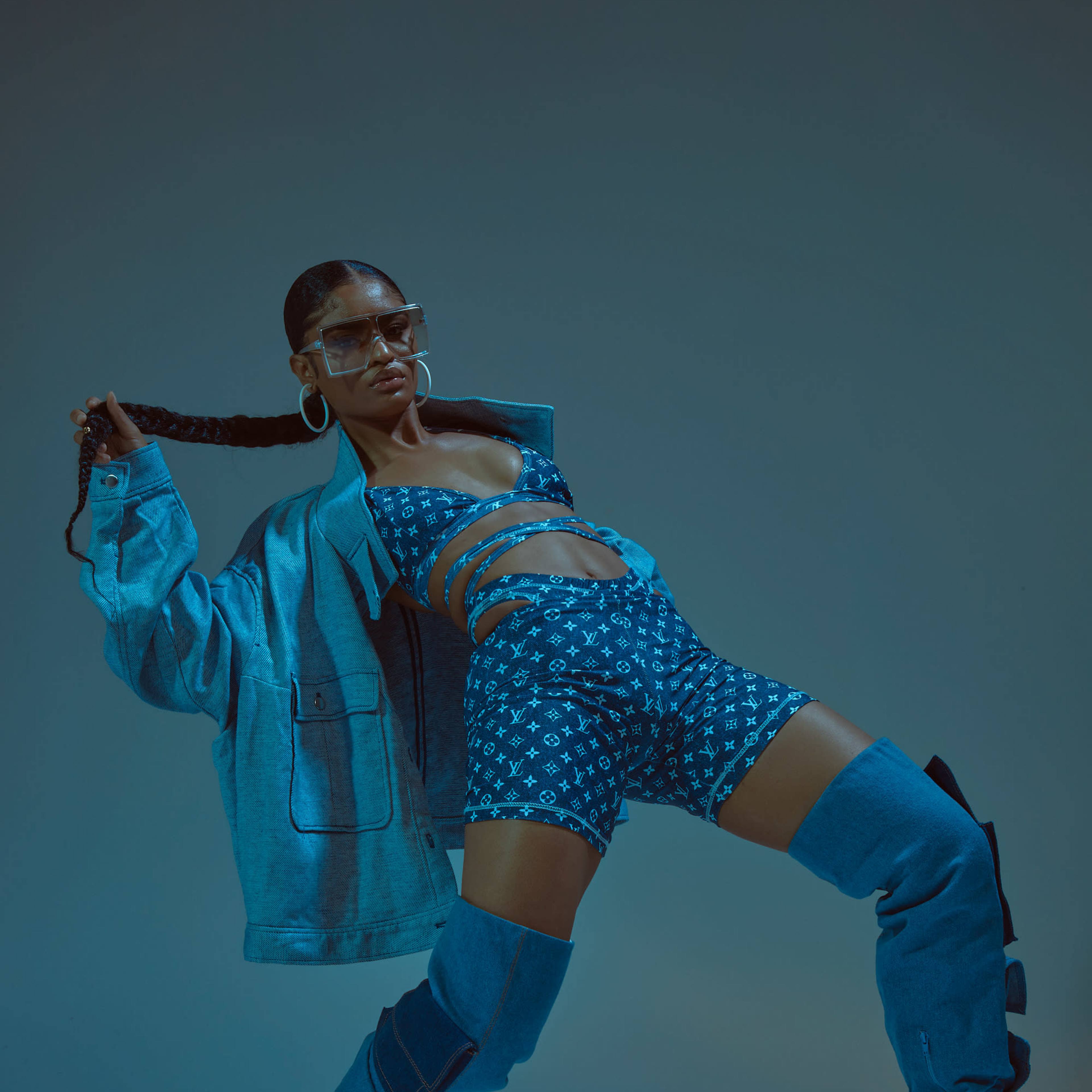 A model poses dynamically in a blue ensemble featuring a cropped top and shorts, complemented by oversized boots and a denim jacket.