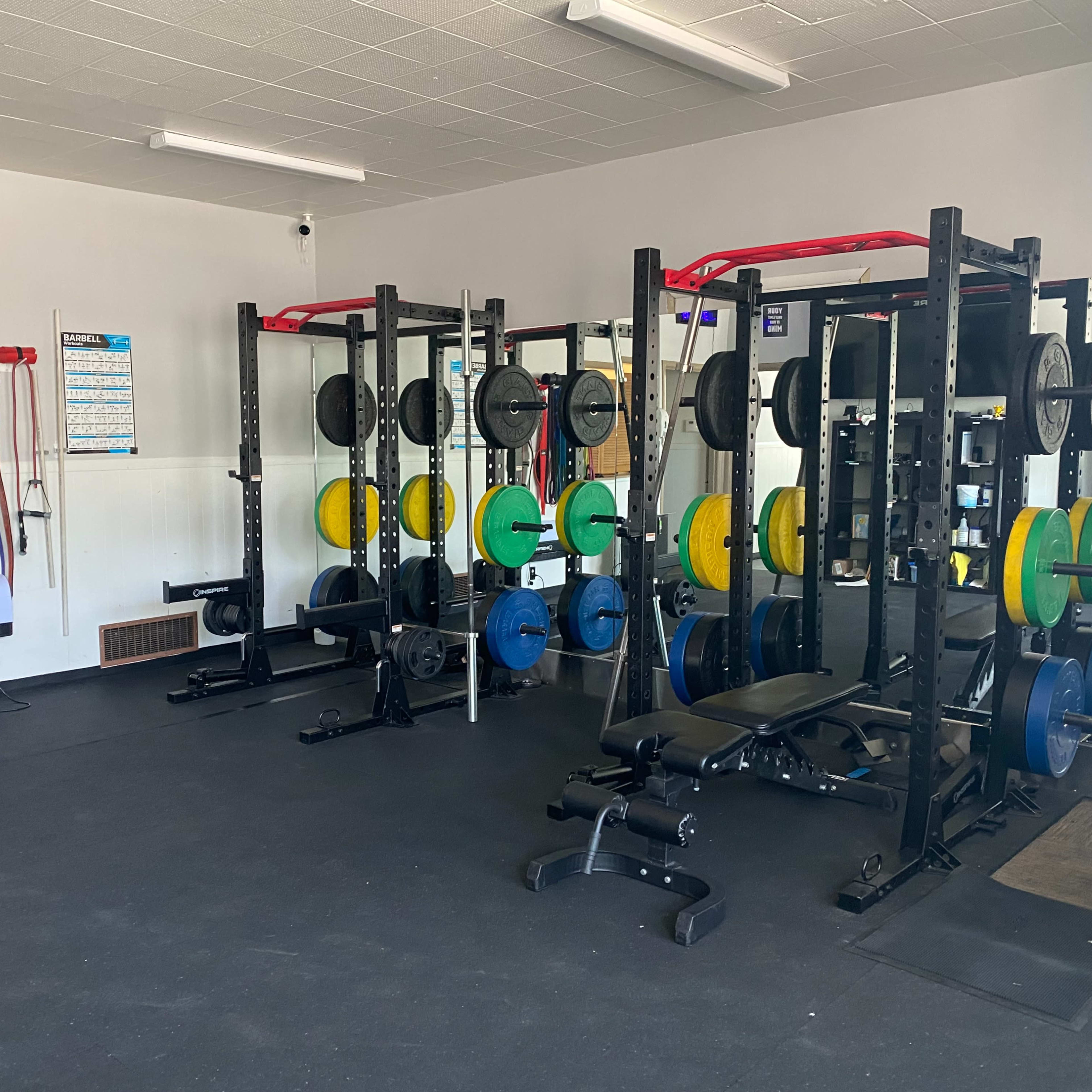 A gym interior featuring multiple weightlifting racks with colorful plates, a bench, and resistance bands hanging on the wall.