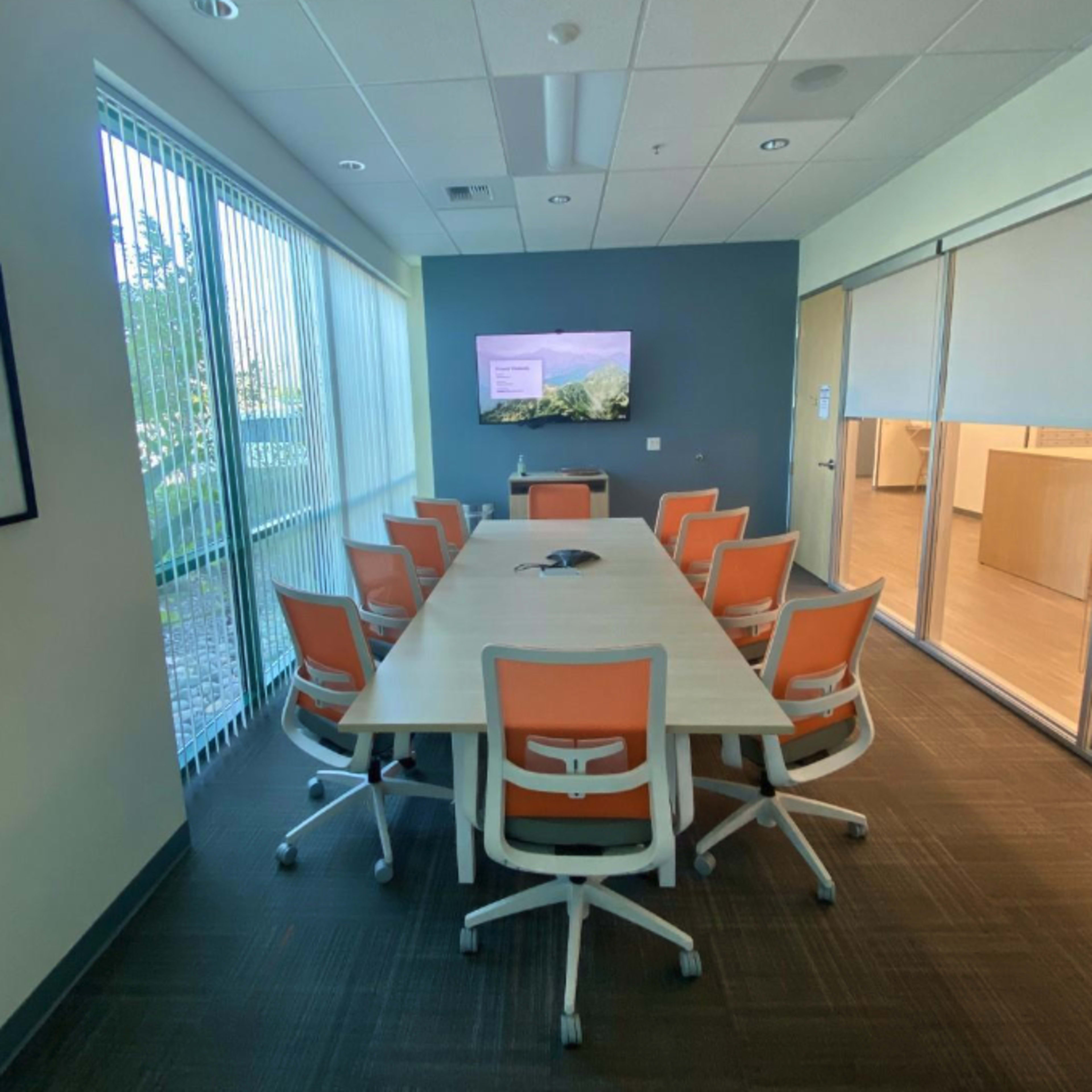 A conference room featuring a long table surrounded by eight orange and white chairs, with a wall-mounted screen displaying a nature image and large windows providing natural light.