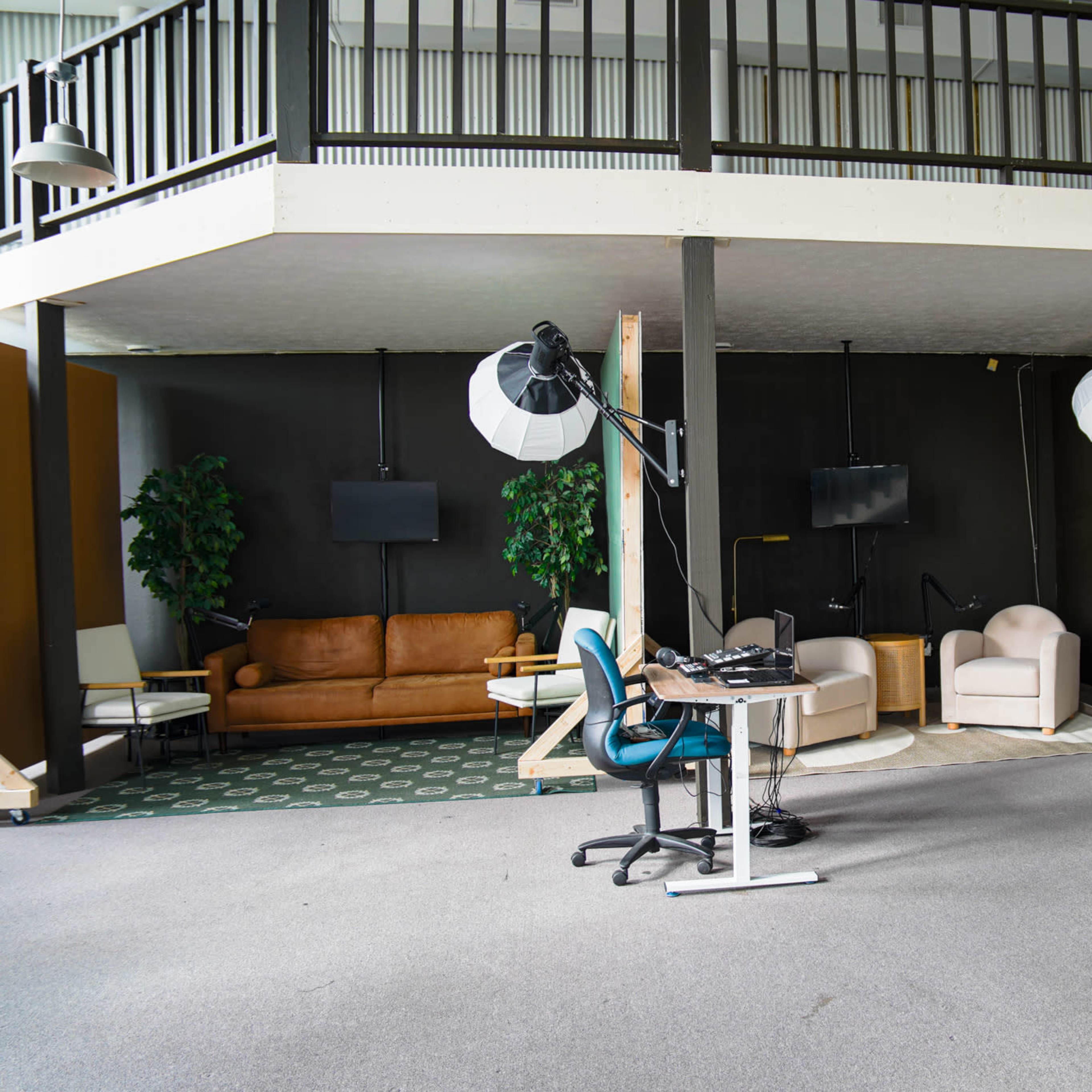 A spacious indoor area with a raised platform, featuring a seating arrangement of a couch and chairs, along with lighting equipment and a desk on the floor below.