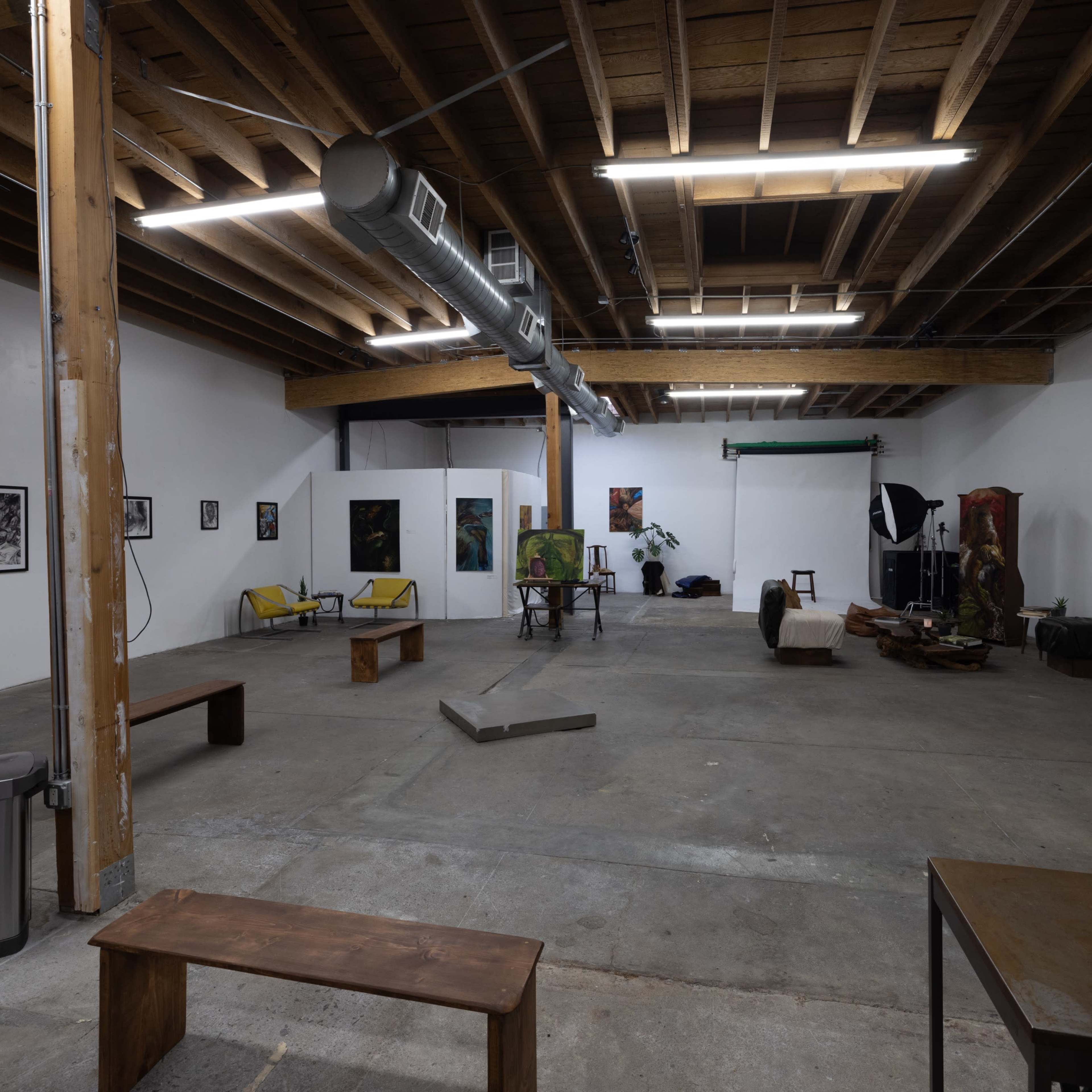 A spacious art gallery with exposed wooden beams, minimal furniture, and various artworks displayed on the white walls.