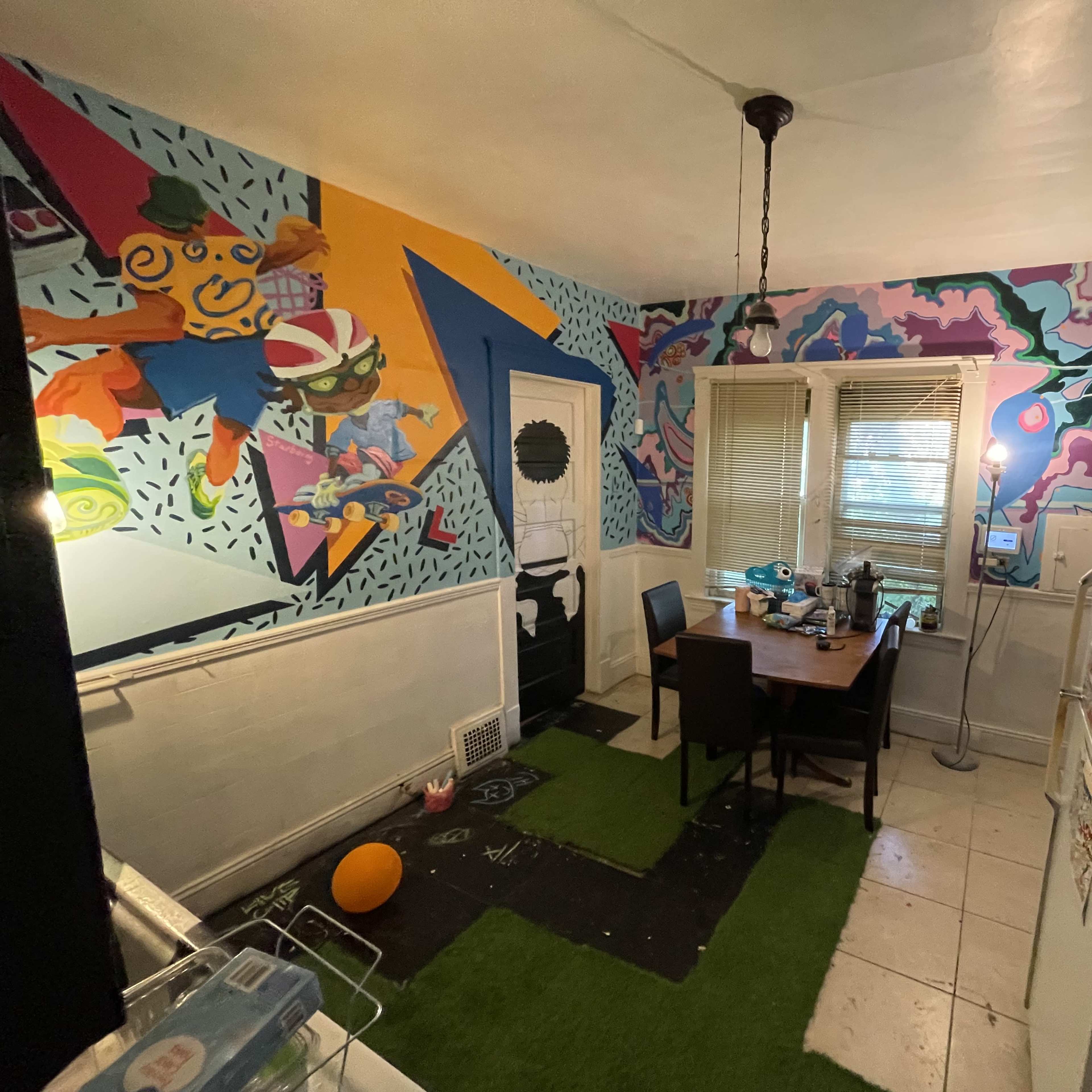 The kitchen features colorful murals on the walls, a table with chairs, and a refrigerator against one side.