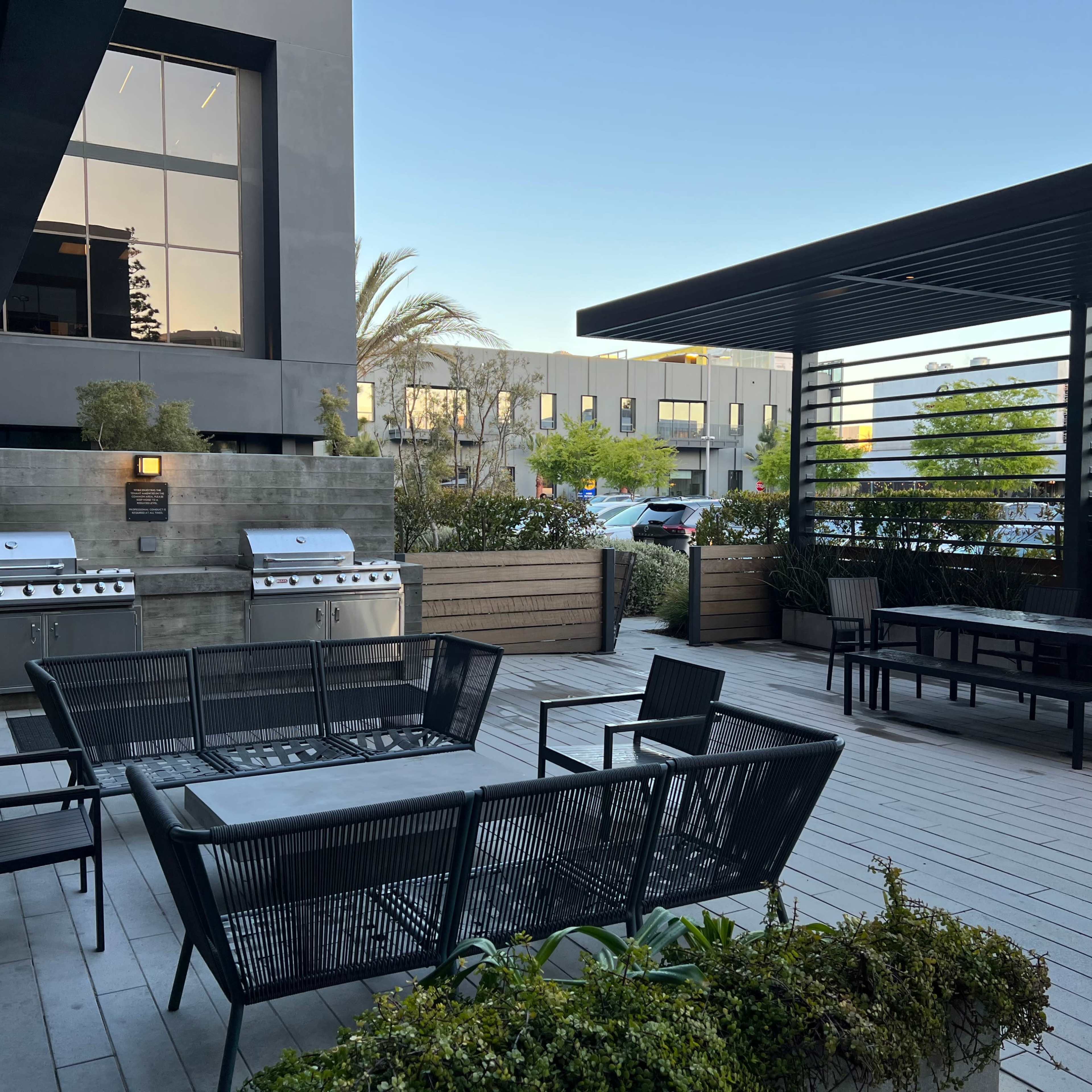 The scene shows a modern outdoor patio area with two barbecue grills, black seating, and tables surrounded by greenery and a building in the background.