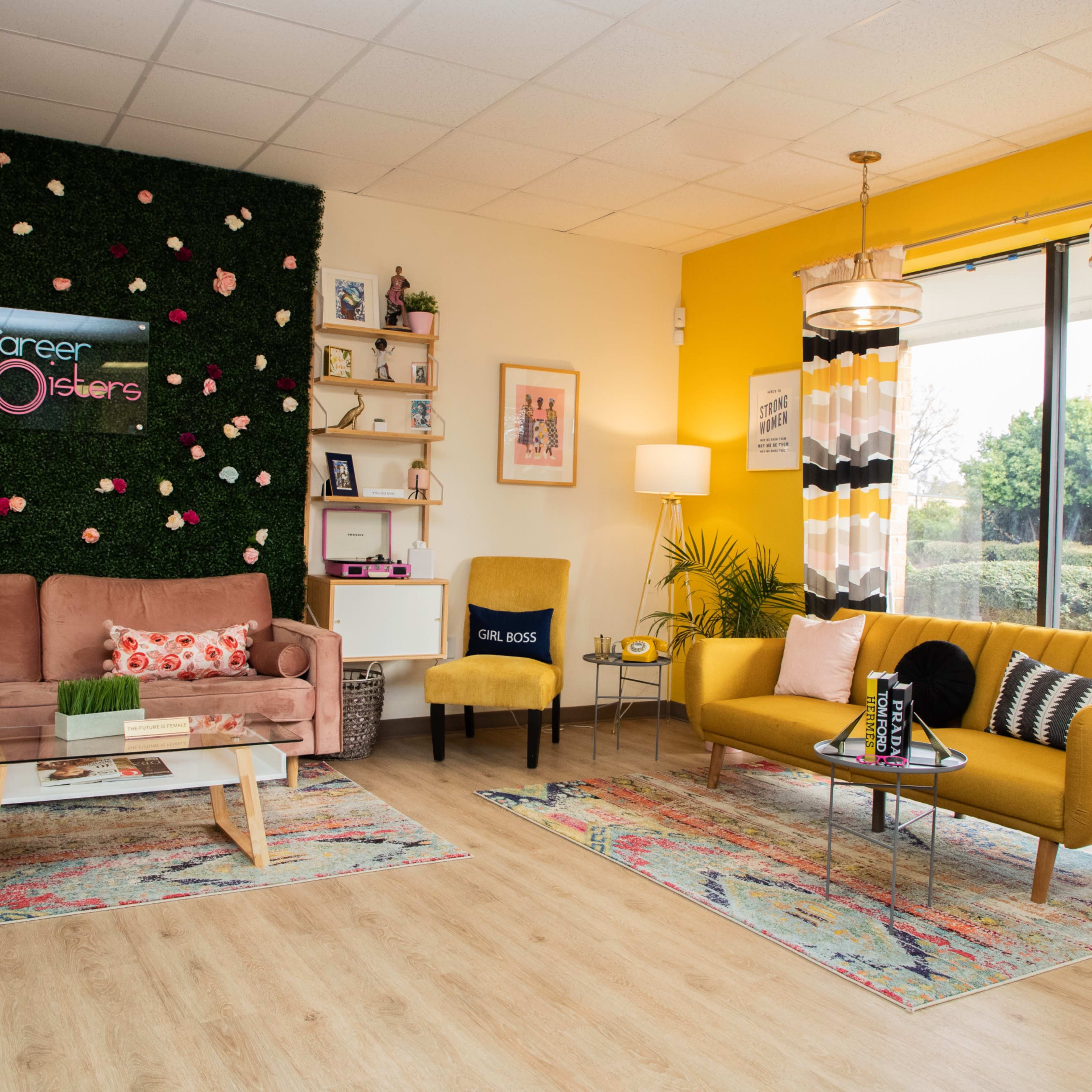 A bright, modern office space featuring a yellow accent wall, a green plant wall, two stylish sofas, and decorative rugs.