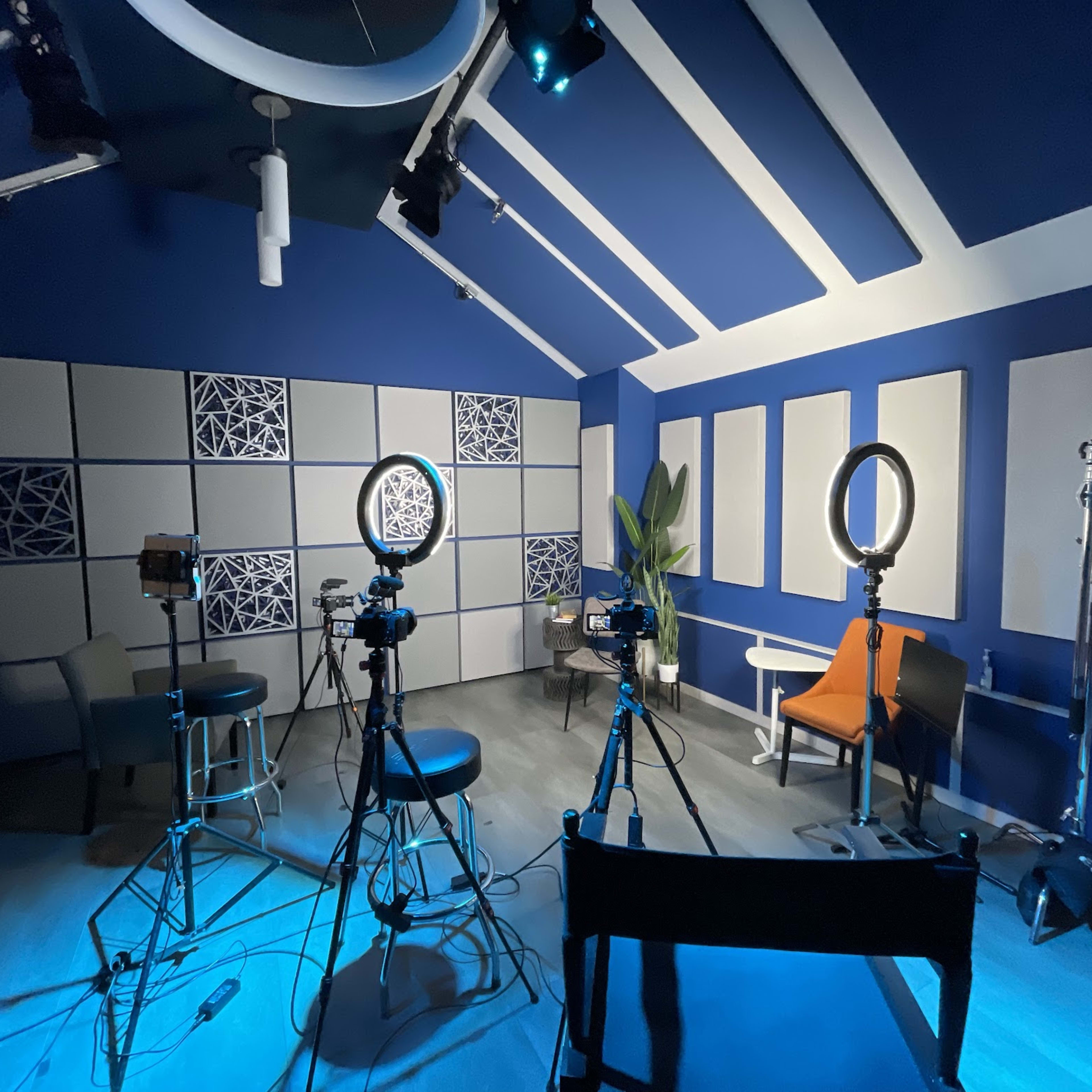 A filming studio with several cameras, lighting equipment, and a few chairs arranged against a blue wall.