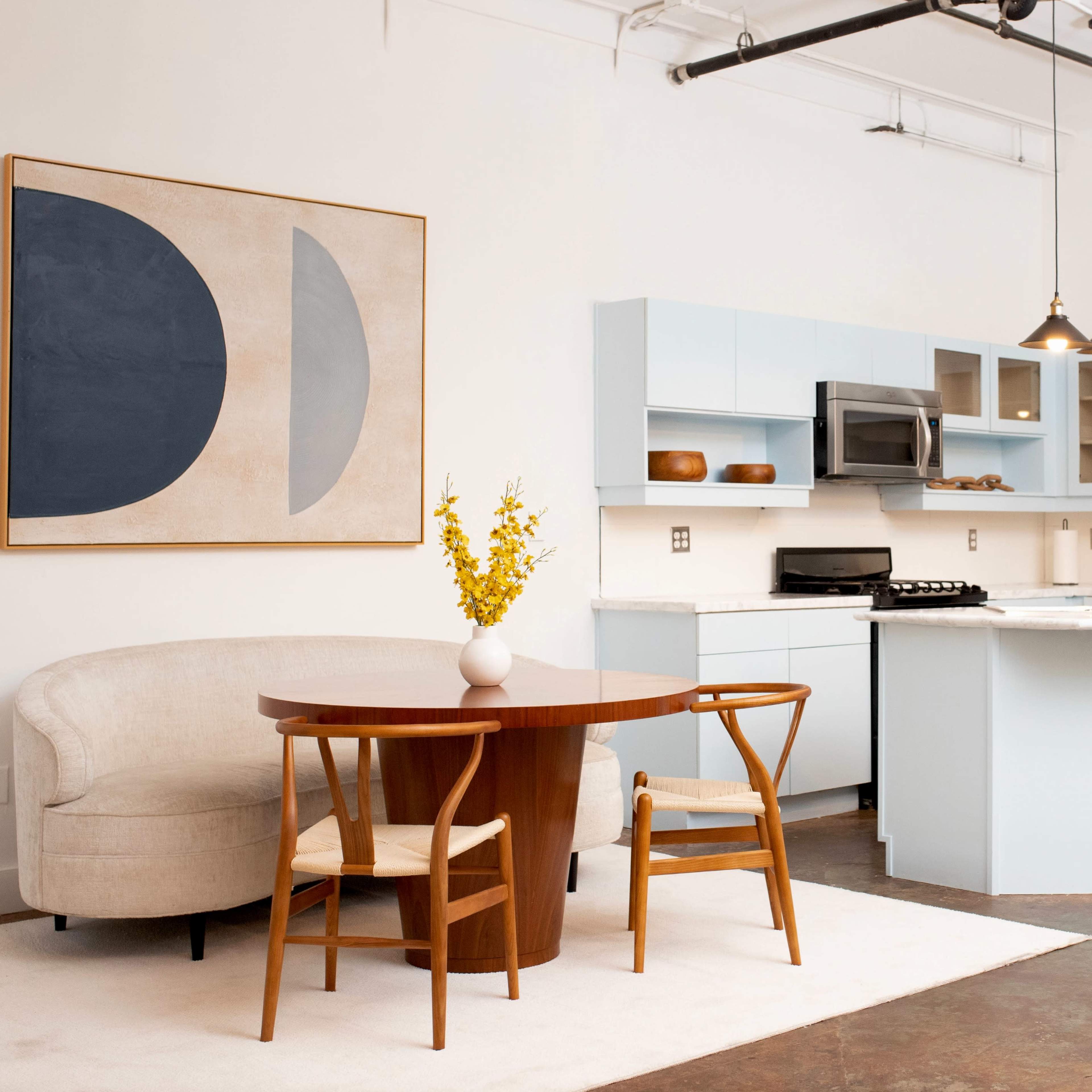 A modern kitchen and dining area feature light blue cabinetry, a round wooden table with two chairs, and a sofa, with a large abstract painting on the wall.
