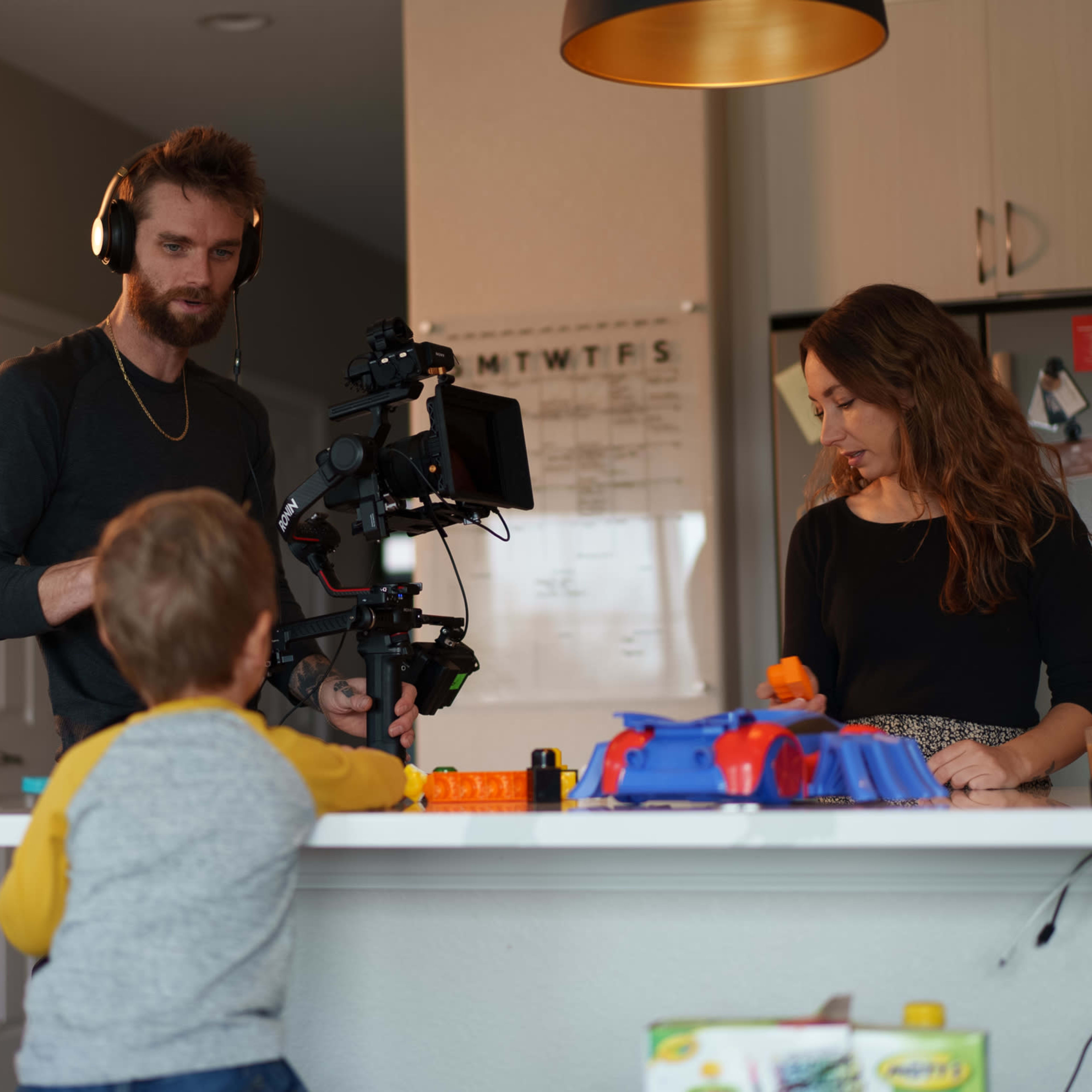 A man with headphones operates a camera on a stabilizer while a woman watches in a modern kitchen, as a child observes from the countertop.