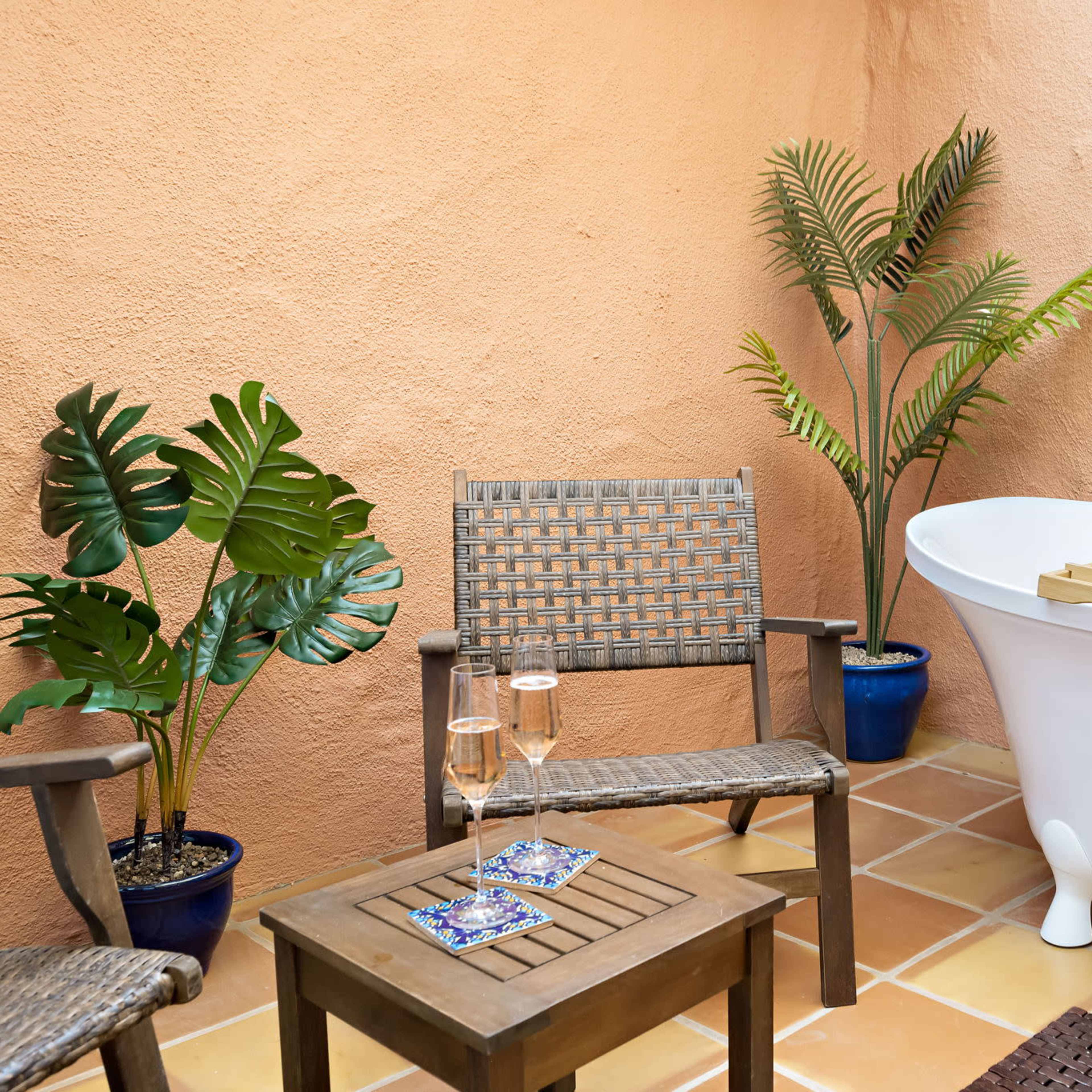 A bathroom setting with a white freestanding bathtub, two wooden chairs, and potted plants against an orange wall.