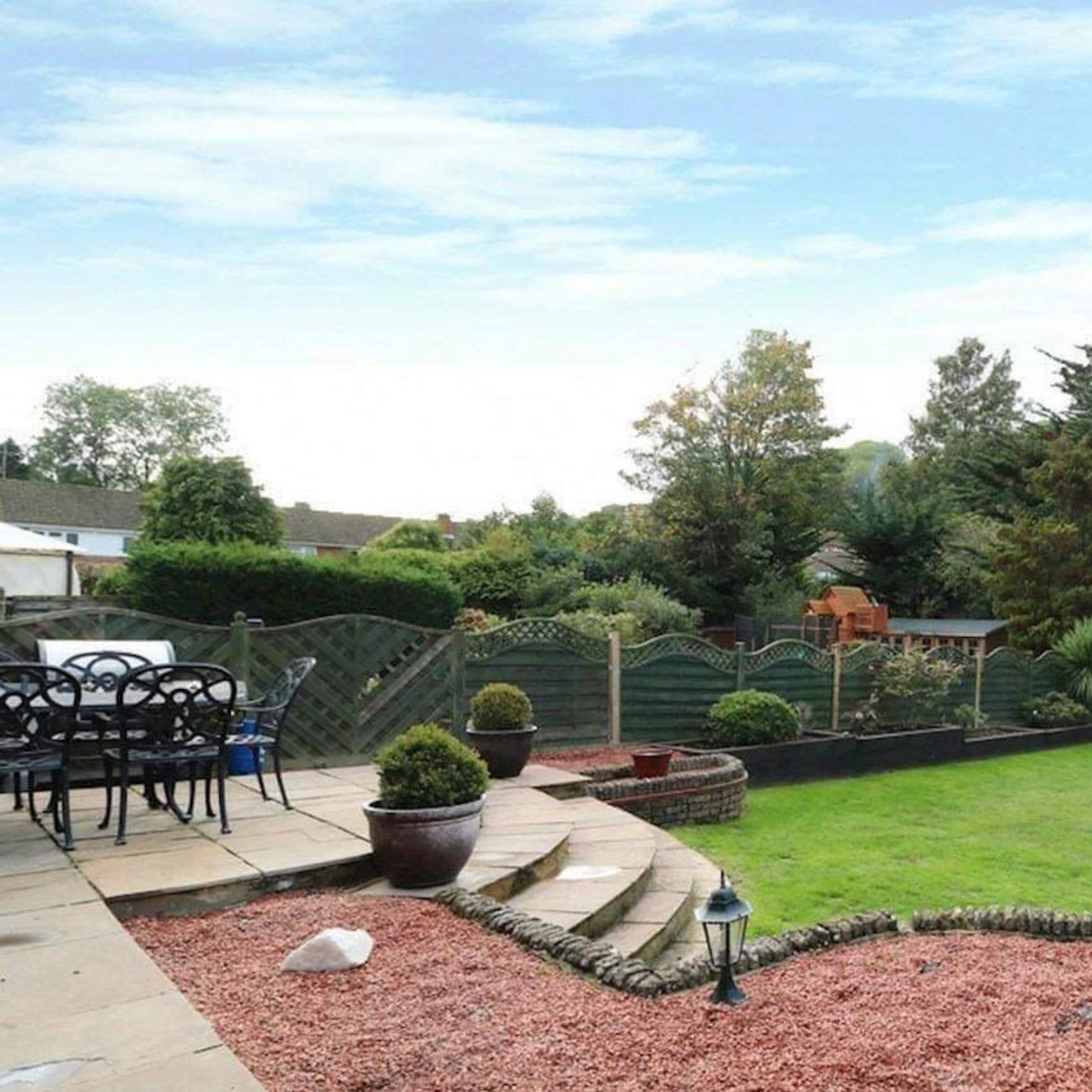 A landscaped backyard featuring a grassy area, a stone patio with a table and chairs, decorative plants, and a playset in the background.