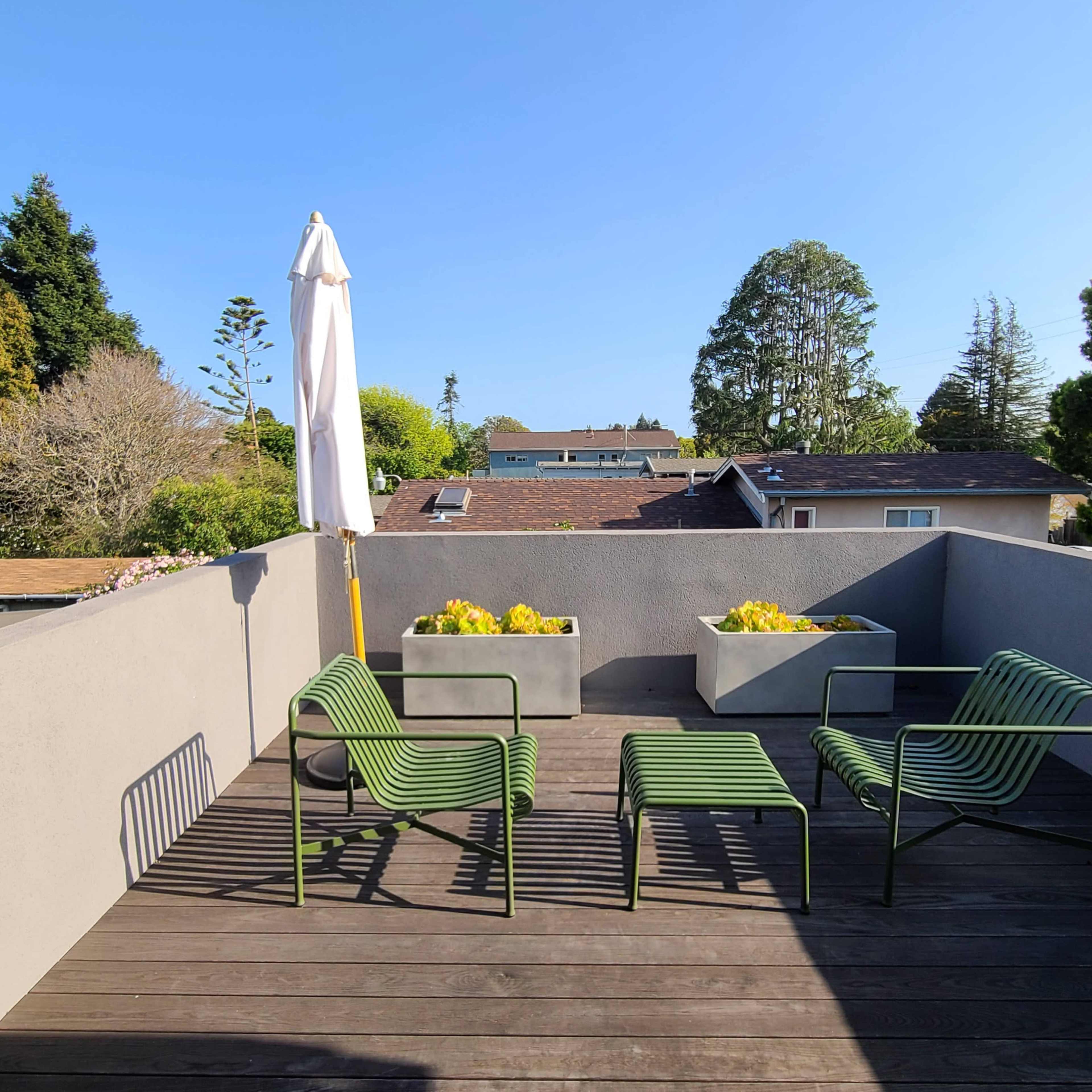 A rooftop terrace with green metal furniture, including lounge chairs and a small table, surrounded by planters with yellow flowers.
