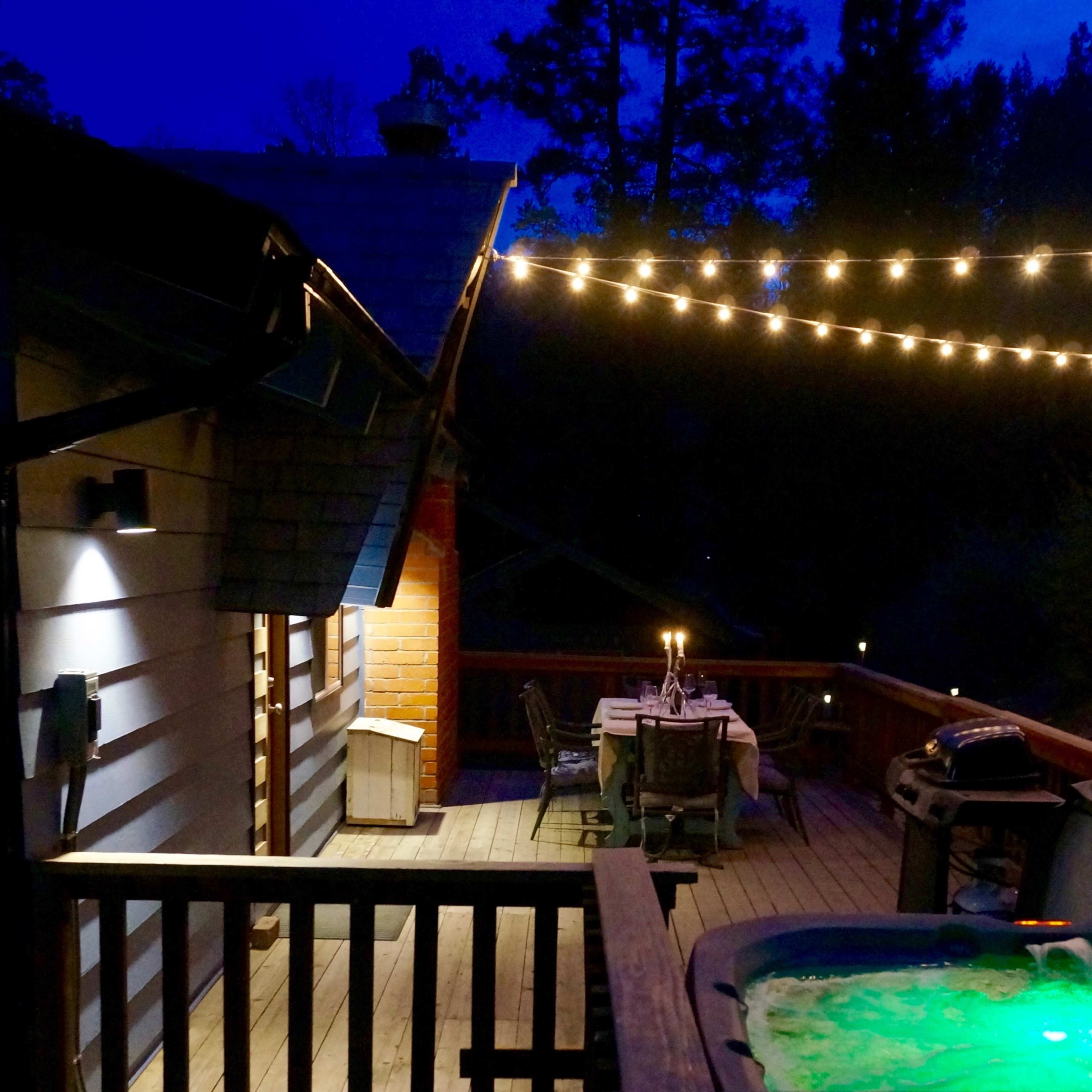 A dimly lit deck with a hot tub and a table set for dining, illuminated by string lights against a twilight sky.