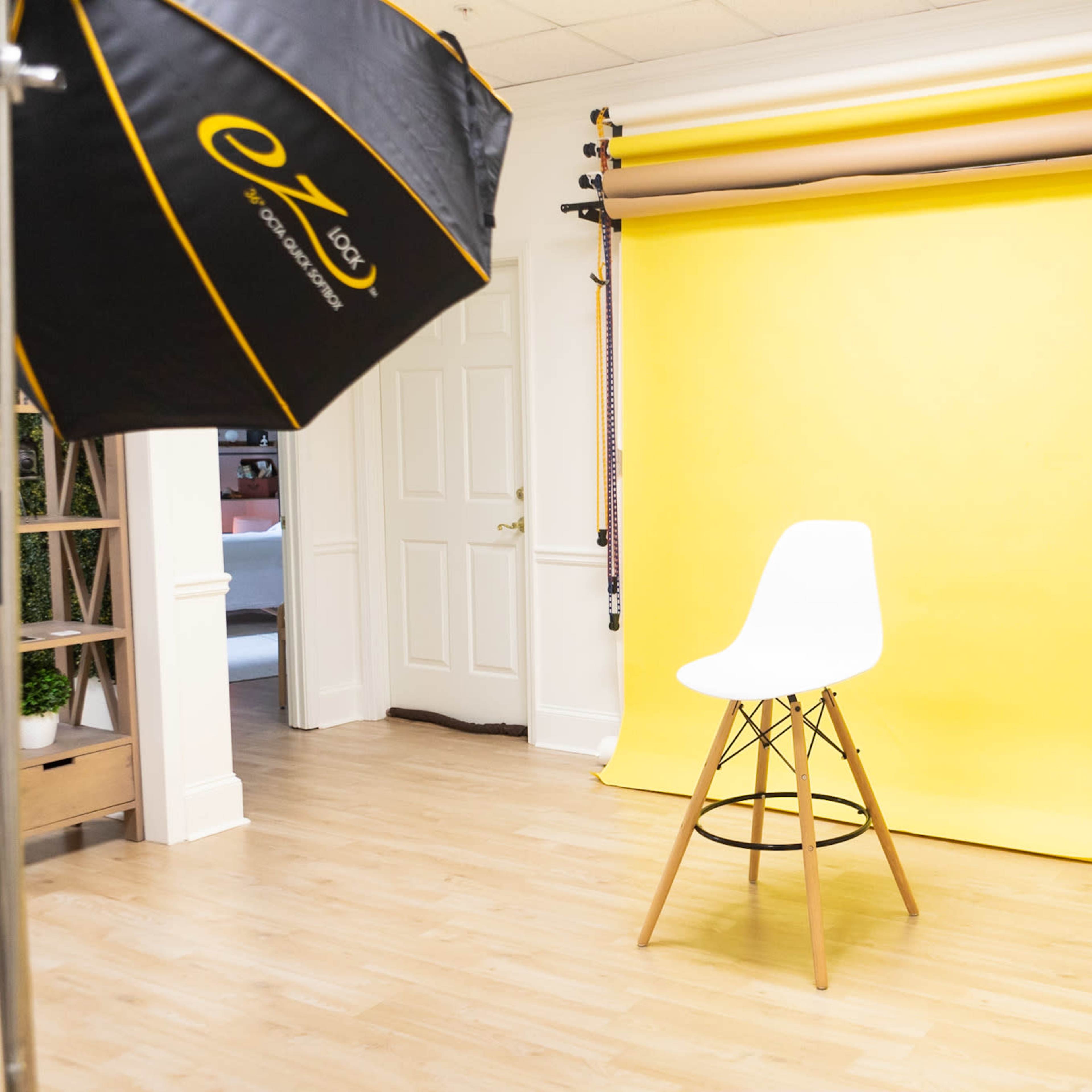 A photography studio with a white chair in front of a bright yellow backdrop, surrounded by lighting equipment and a neutral floor.