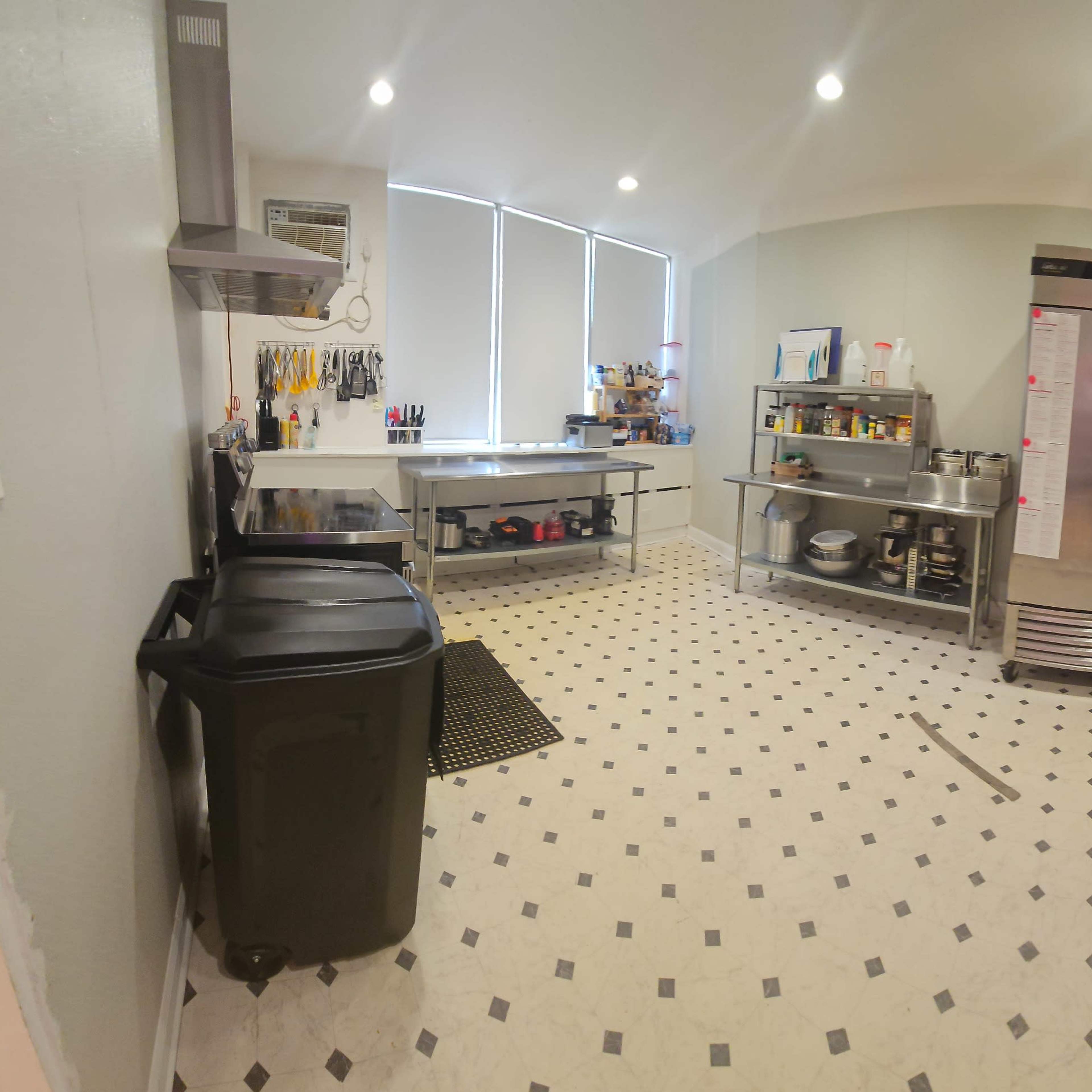 A spacious commercial kitchen with stainless steel appliances, worktables, and a storage area featuring racks and a doorway leading to another room.