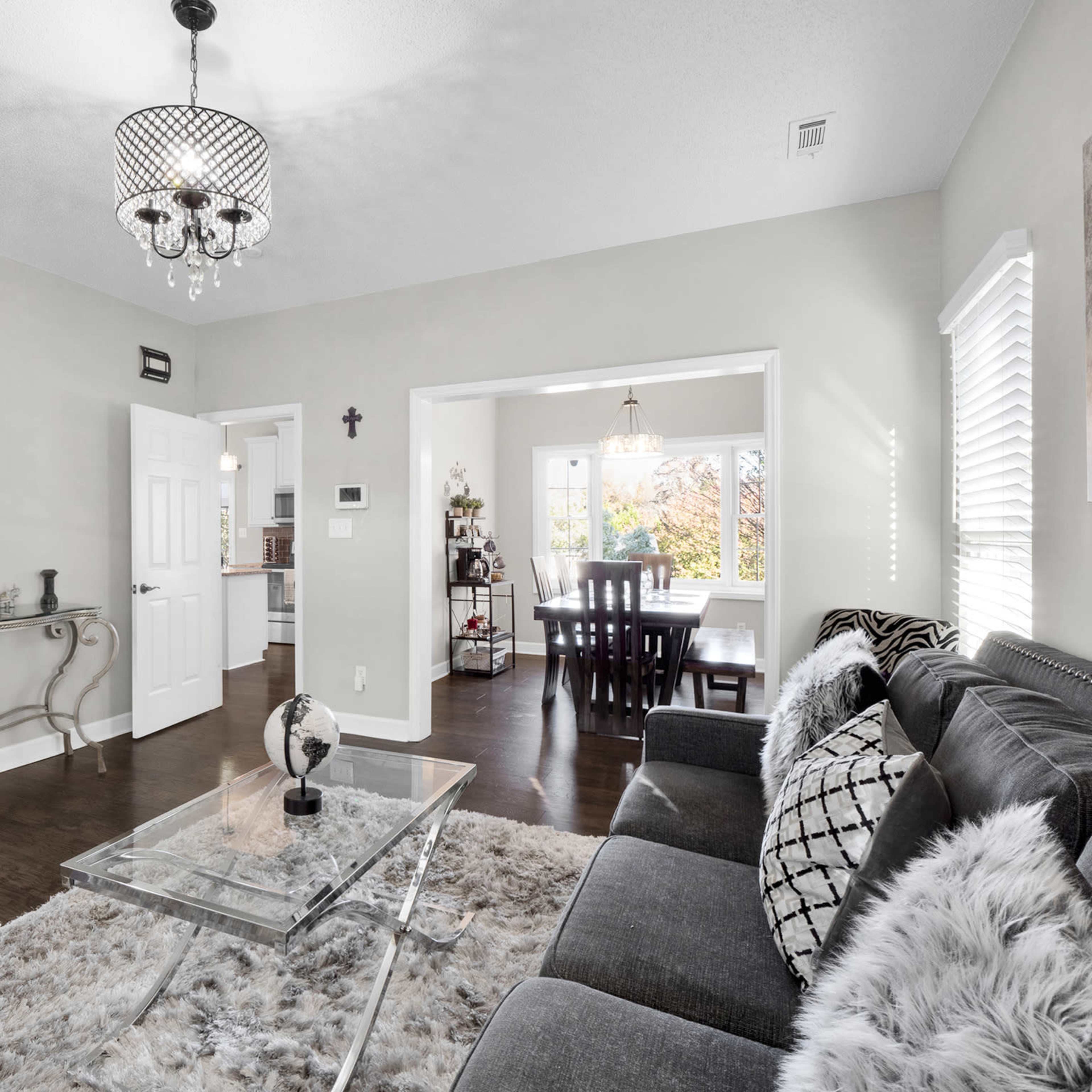 A well-decorated living room with a gray sofa, a glass coffee table on a plush rug, and an adjoining dining area visible through an open doorway.