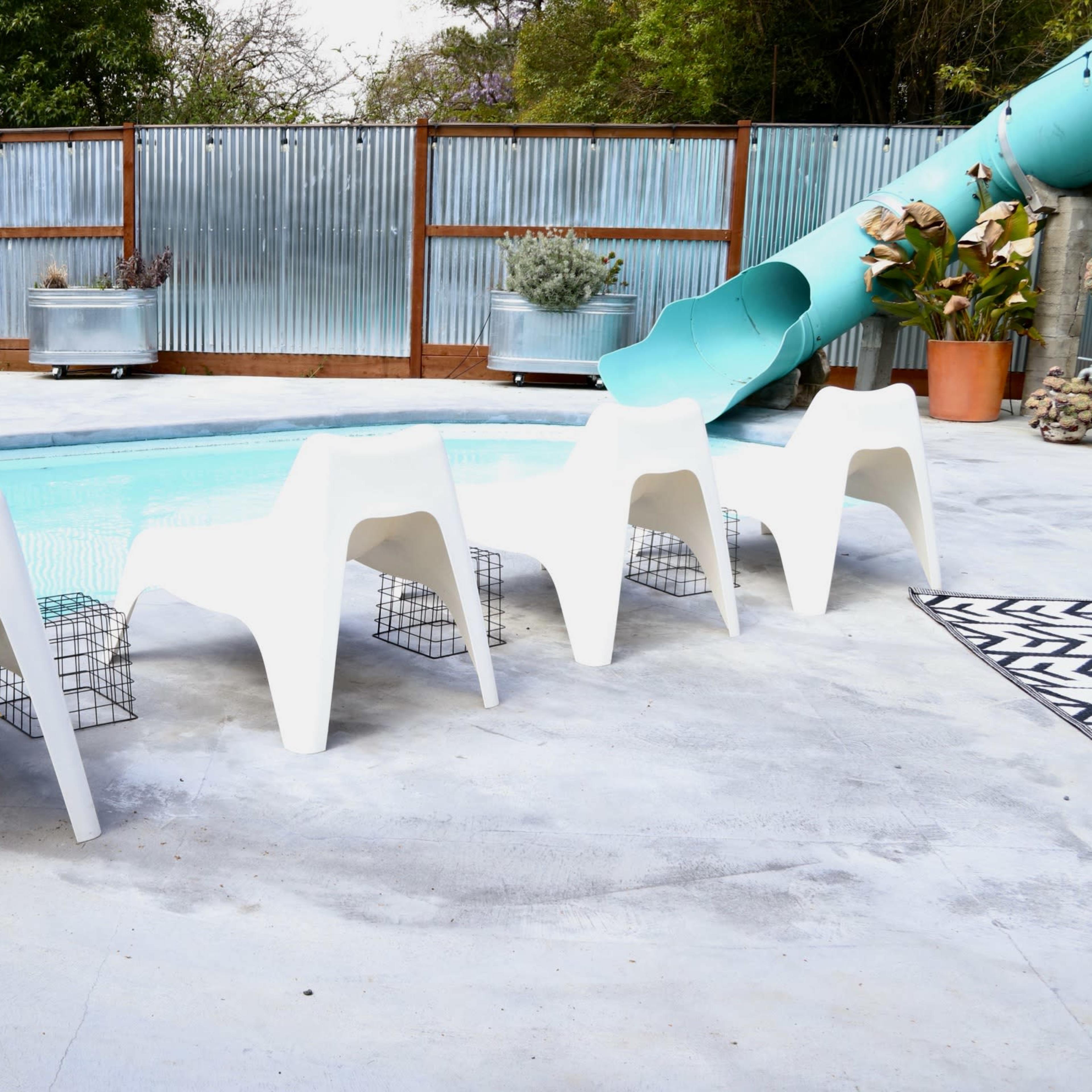 The scene features a swimming pool surrounded by four white lounge chairs, two wooden deck chairs, and a green water slide in a yard with corrugated metal fencing.