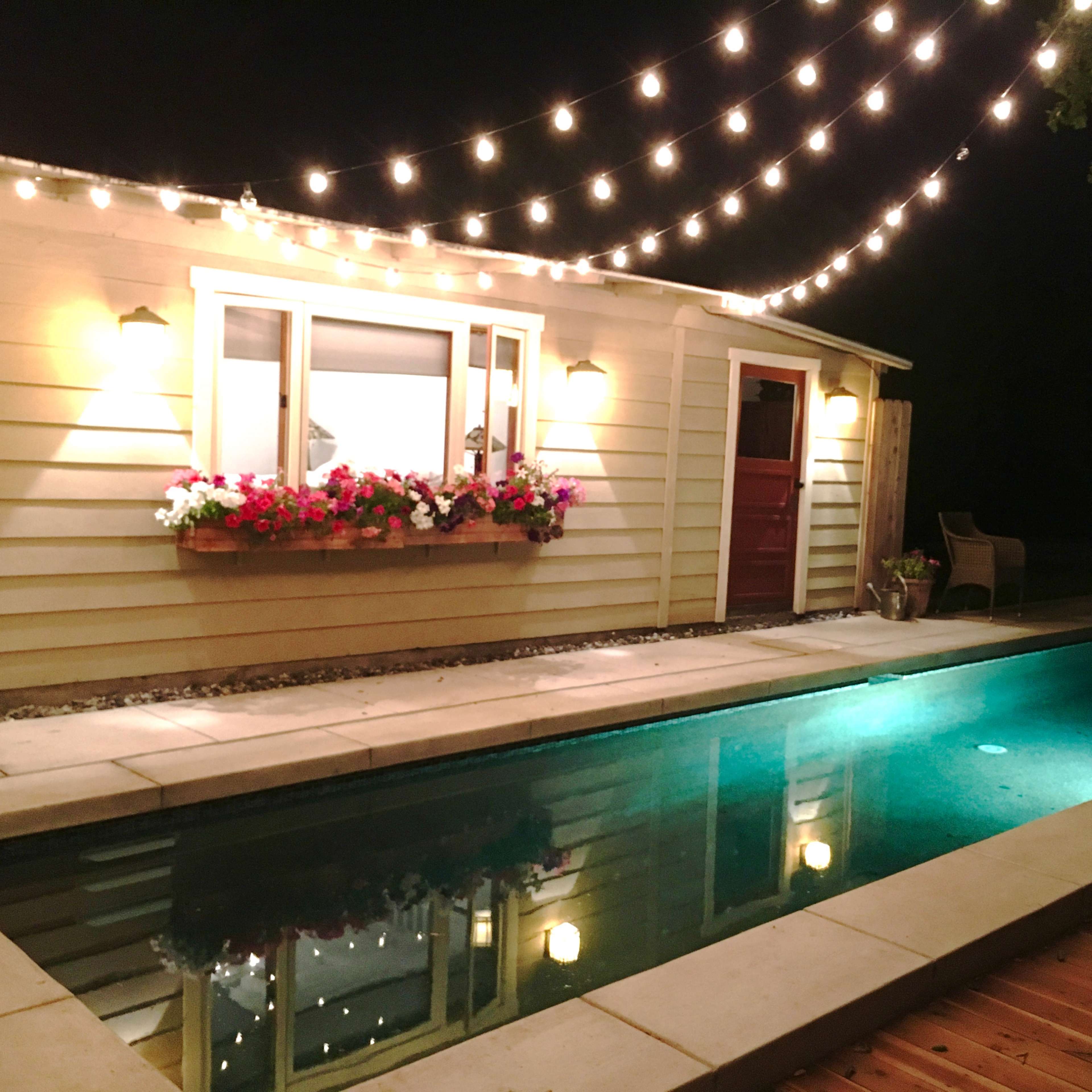 A well-lit pool area features a quaint building with flower boxes under the windows, surrounded by string lights at night.