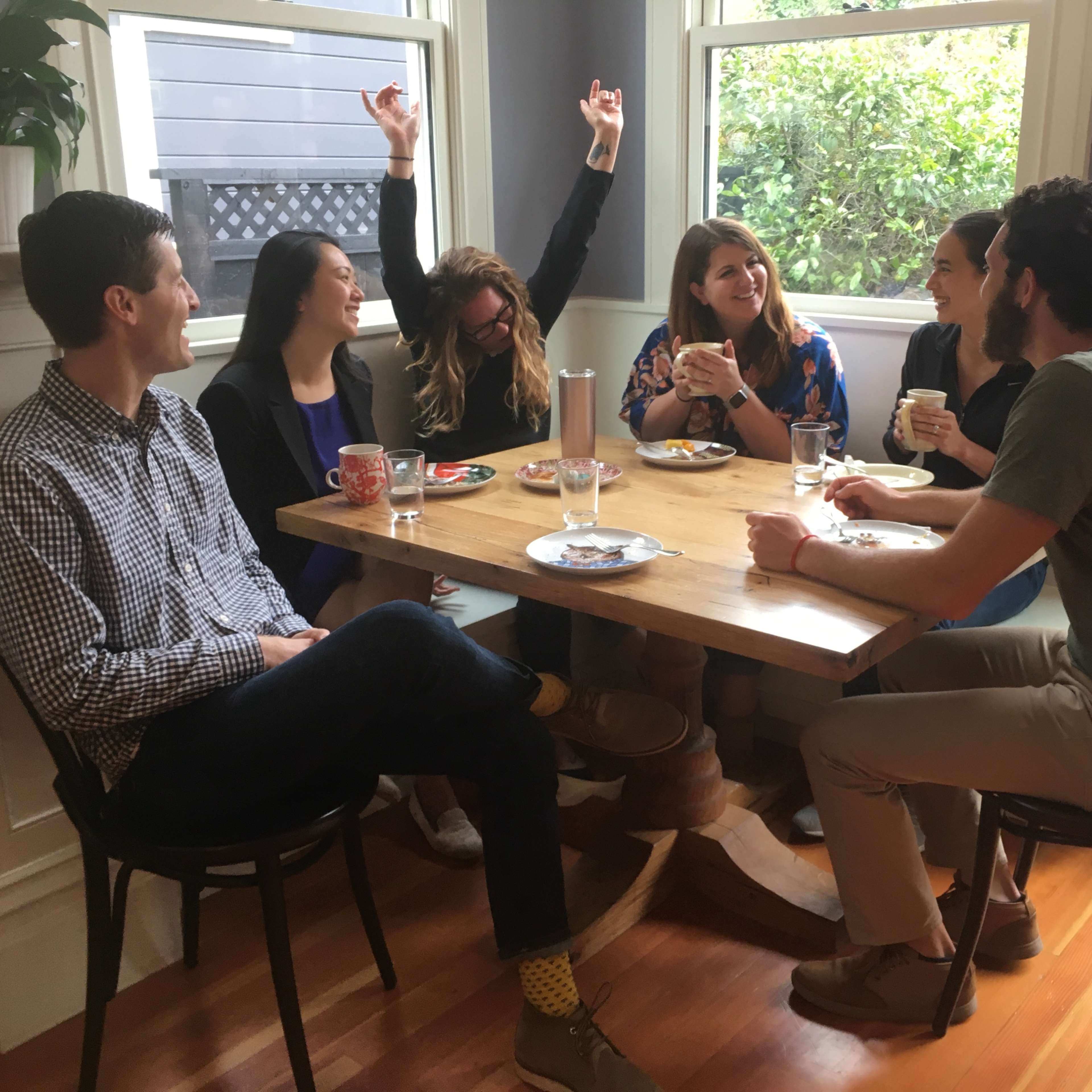 A group of six people sits around a table in a well-lit room, engaging in conversation while enjoying food and drinks.