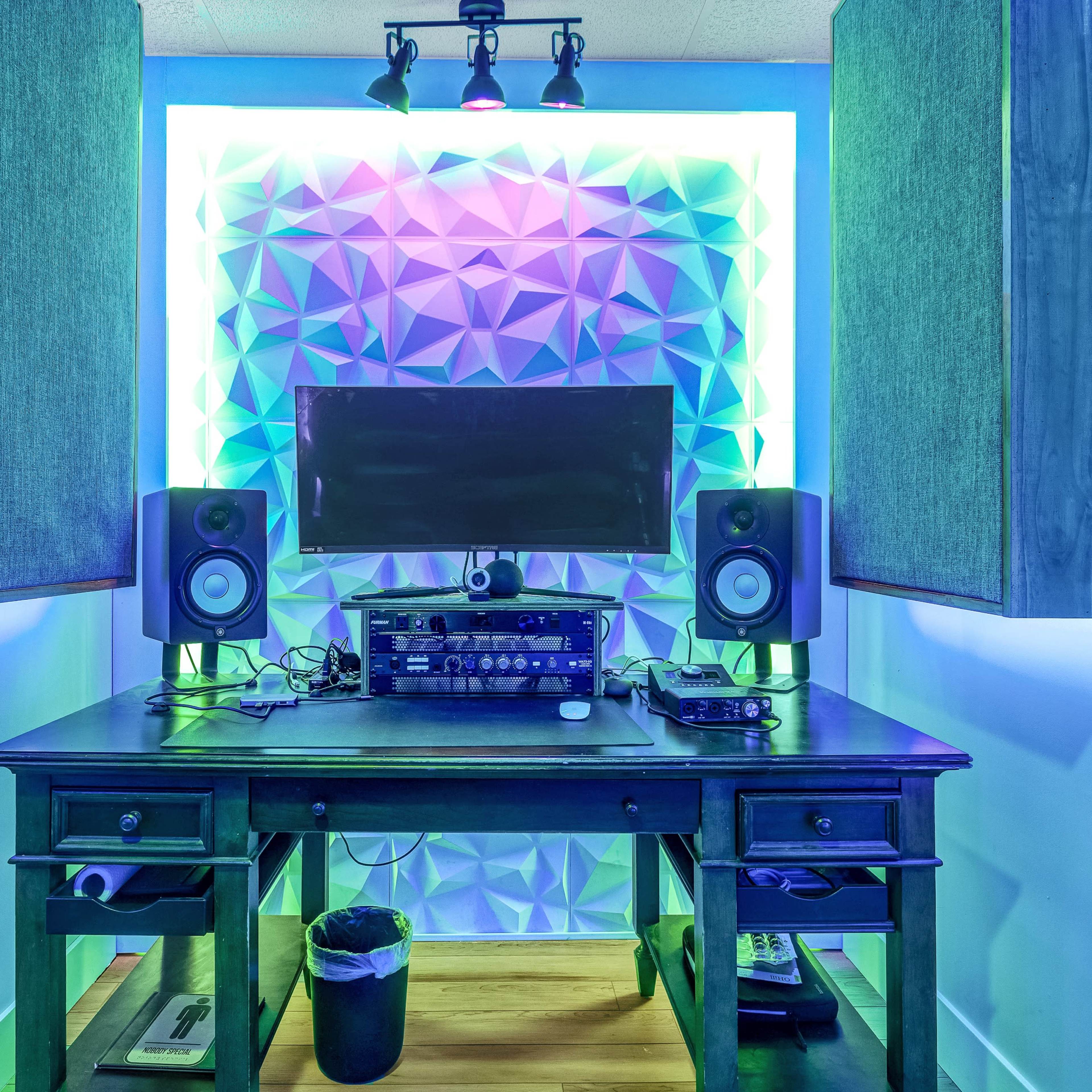 A home recording studio featuring a desk with audio equipment, two speakers, and a monitor, illuminated by colored LED lights against a textured wall.