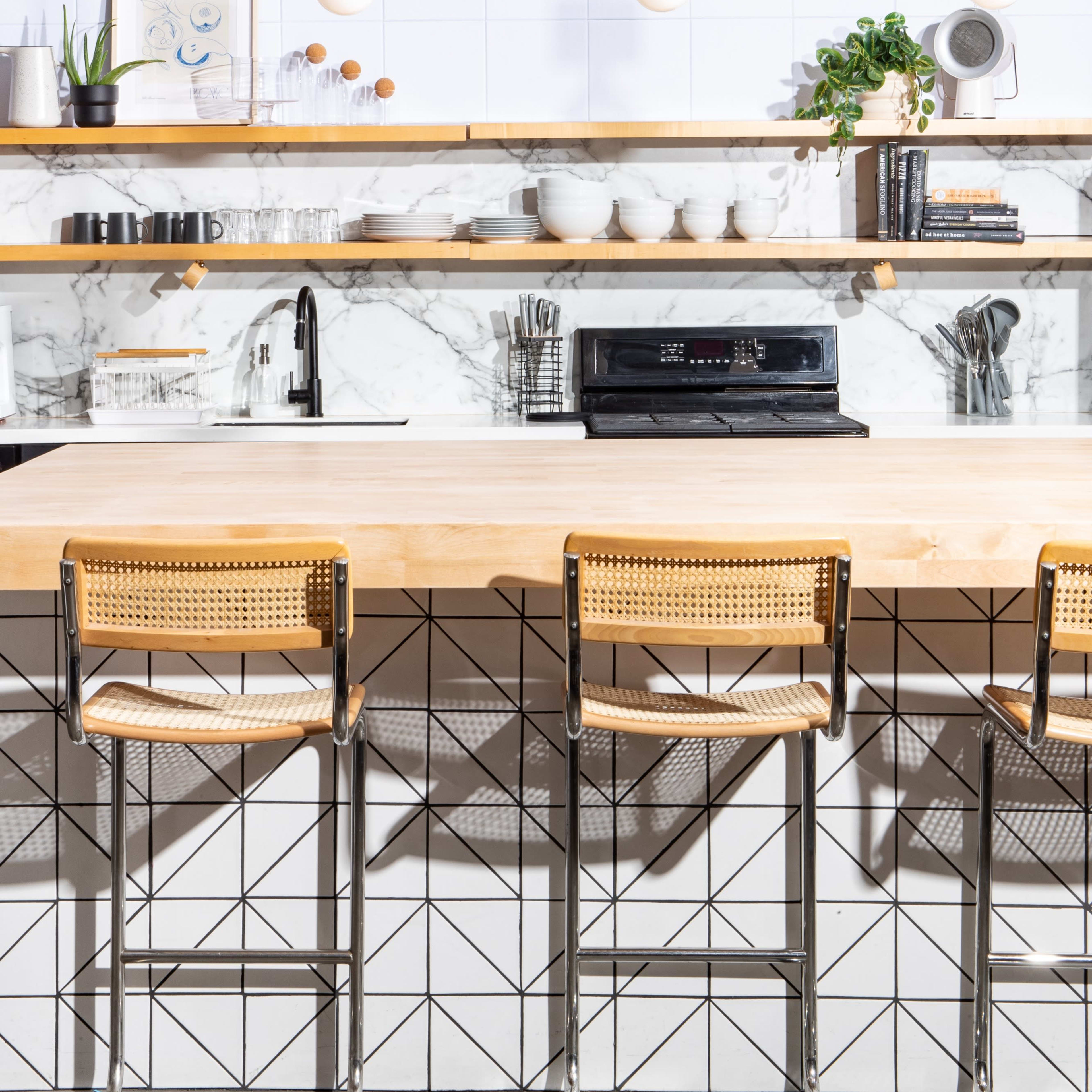 A modern kitchen with a wooden countertop, three bar stools, and a marble backsplash featuring shelves with dishes and decorative items.